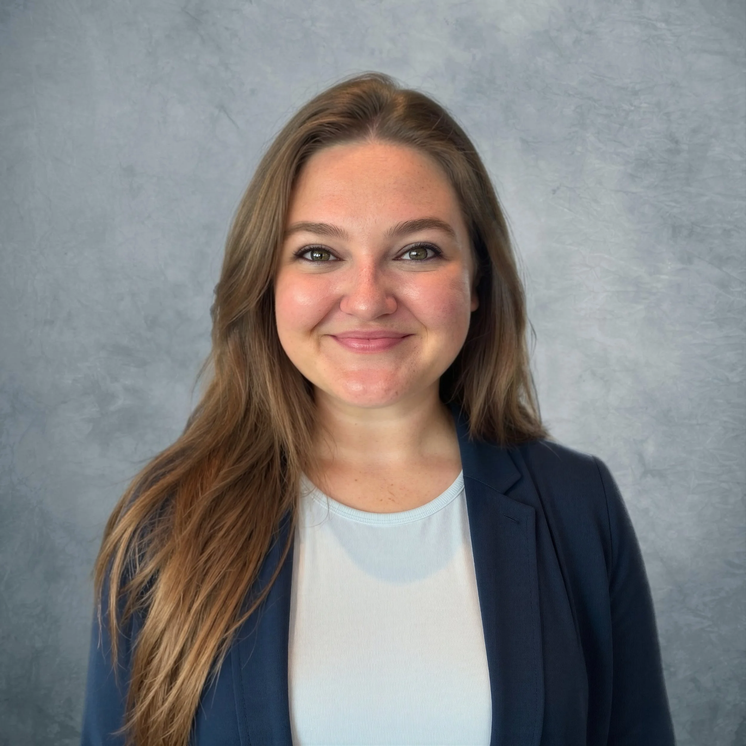 Close-up of a smiling woman with long brown hair, wearing a white top and a dark blazer, against a gray textured background.