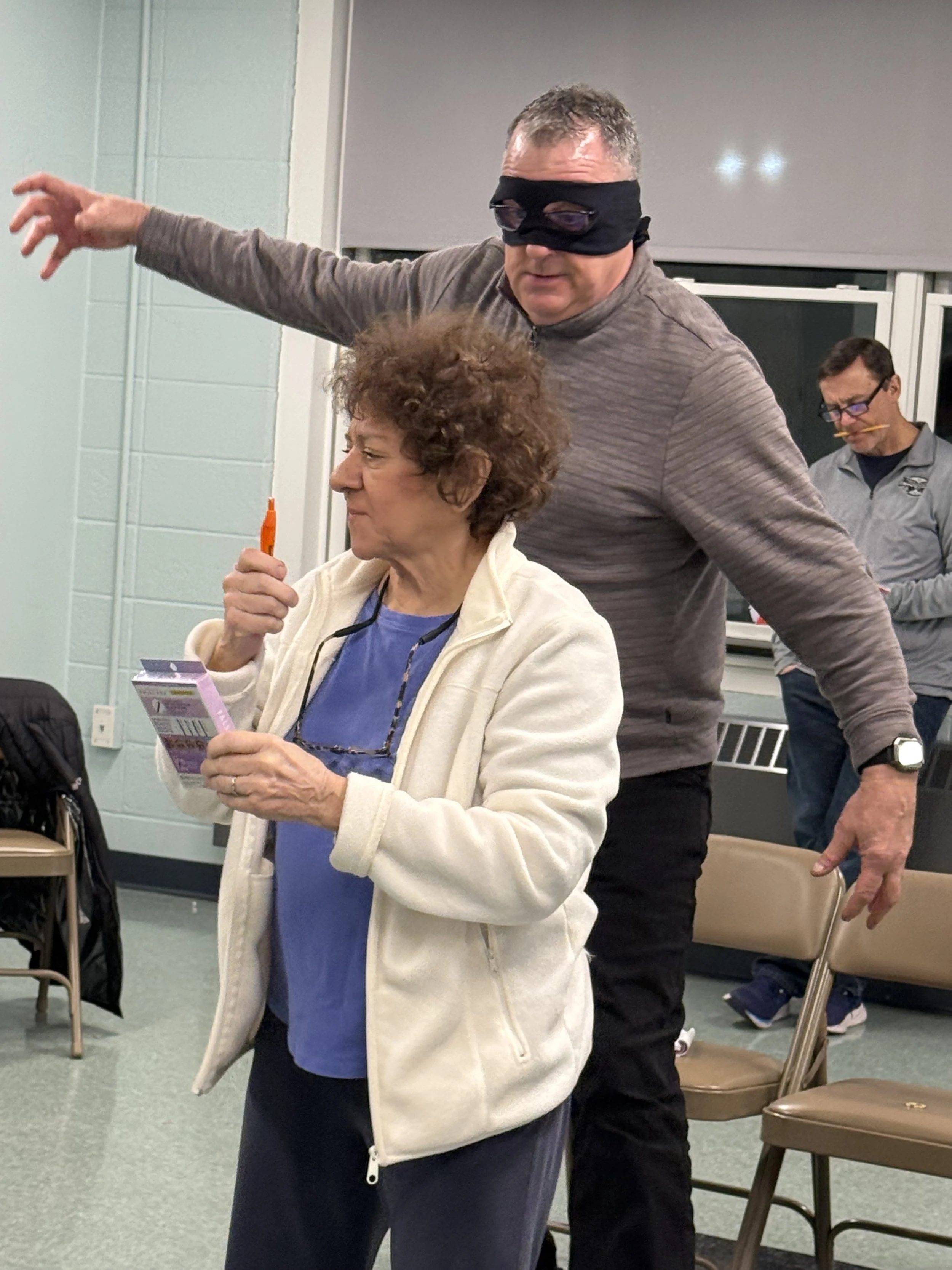 A woman holding a carton of milk, with eyeglasses hanging around her neck and a beige jacket. Sitting behind her is a man with short curly hair, wearing a gray jacket and a black eye mask. In the background, there is another man with glasses, a gray jacket, and a yellow pencil in his mouth. The setting appears to be an indoor meeting or event with chairs, a window, and a pale green wall.