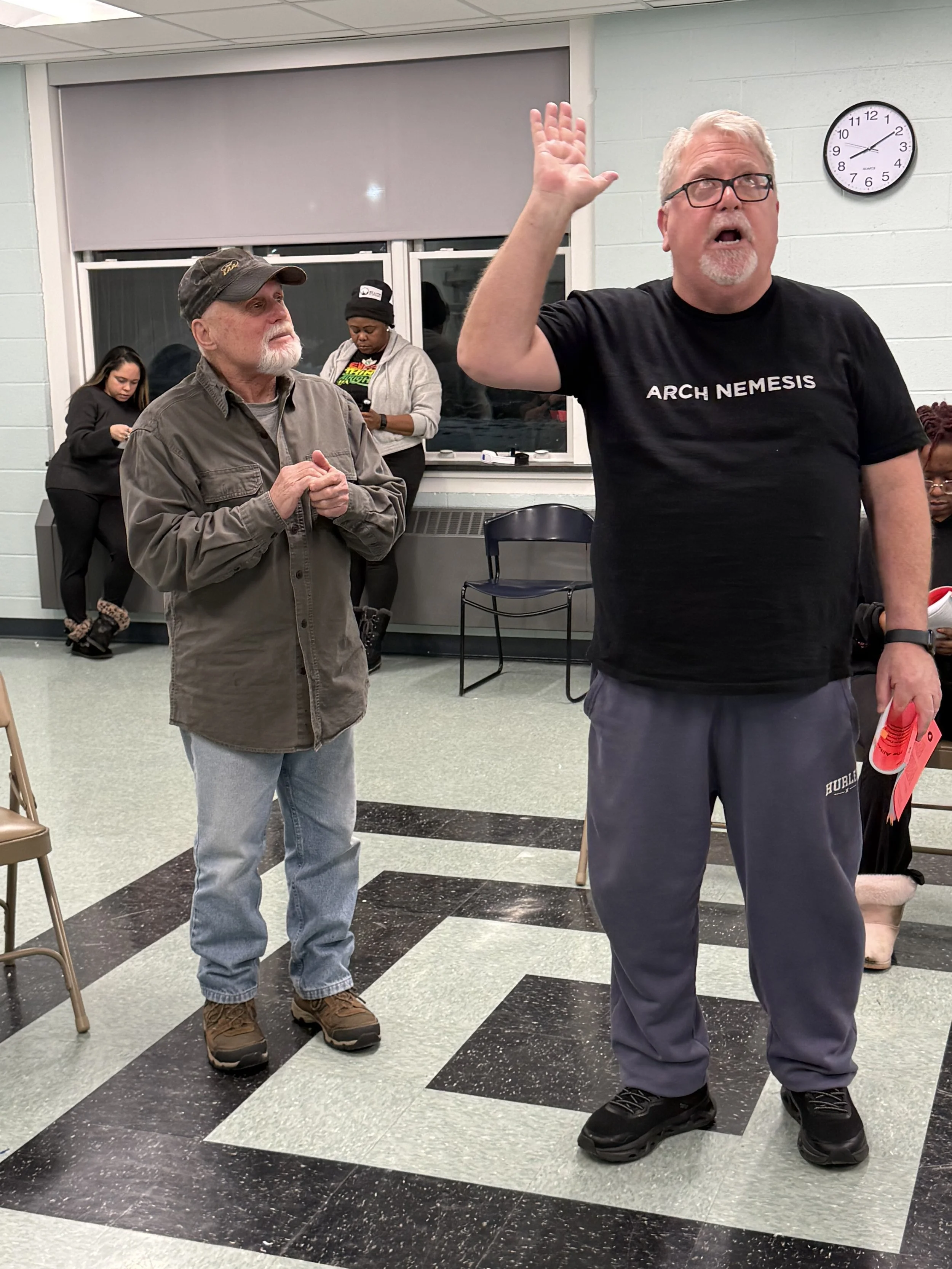 A group of four people inside a room with black and white checkered flooring. Two men are in the foreground, one wearing glasses and a black shirt that says 'Arch Nemesis,' raised hand, and holding a red item. The other man is older, with a gray beard and wearing a cap and gray jacket, looking at the man with glasses. Two women are in the background, one looking at her phone and the other bending over near the window.