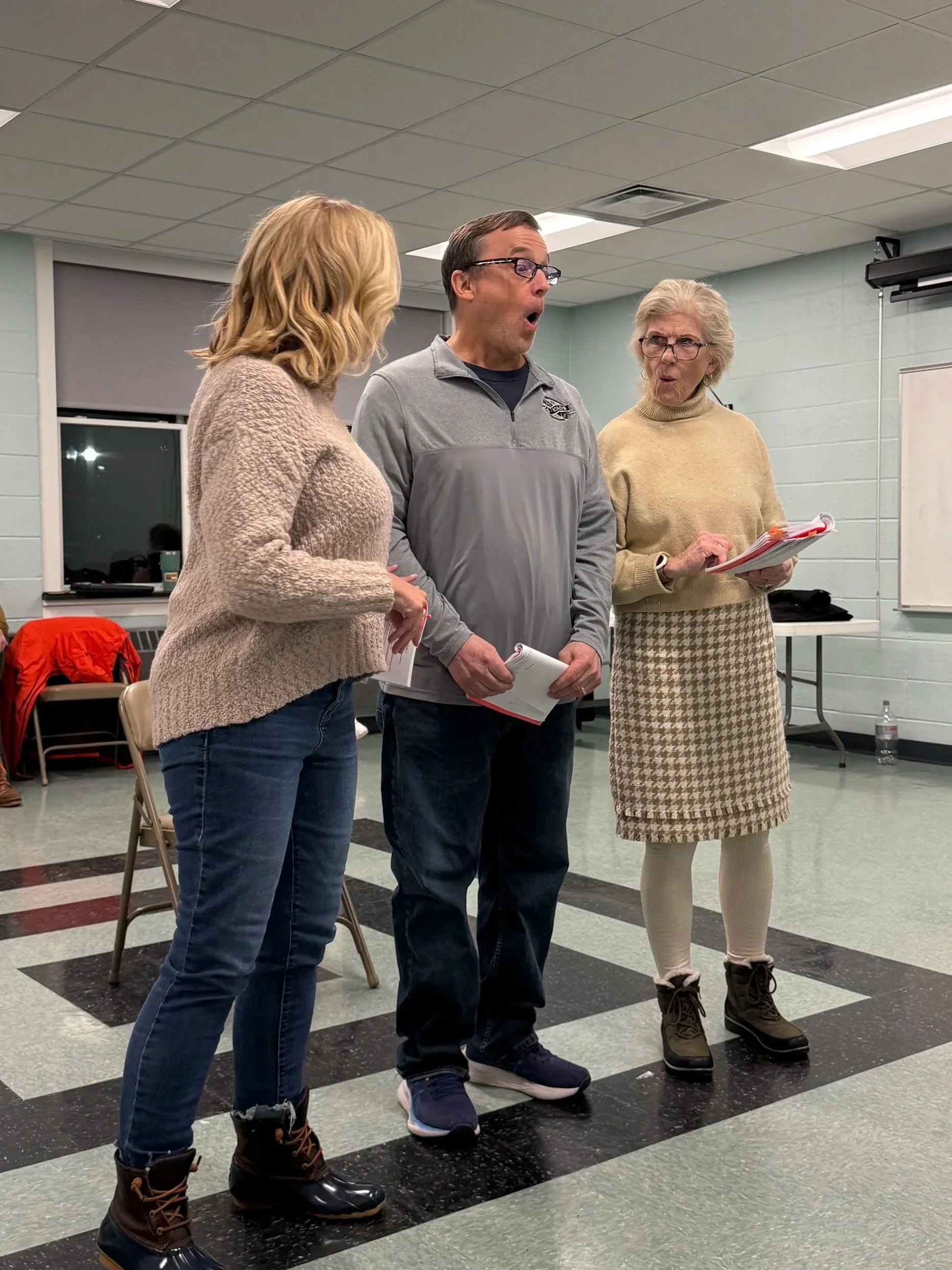 Three people standing in a room, engaged in conversation. The person in the center appears surprised or amazed. Two women and one man, all holding papers or notebooks.
