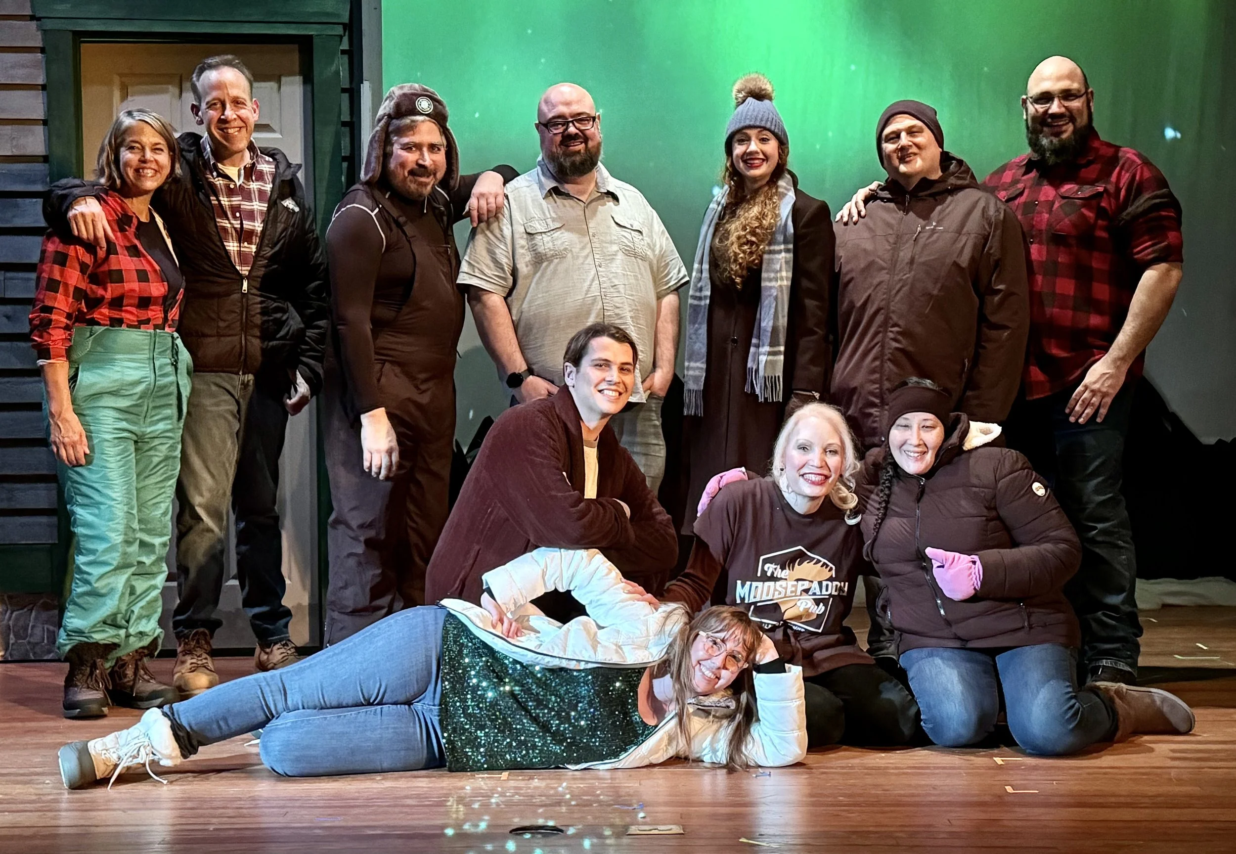 A group of people on a stage, some standing and some sitting or lying down, smiling and posing for the photo. The background is green, and the stage has a wooden floor.