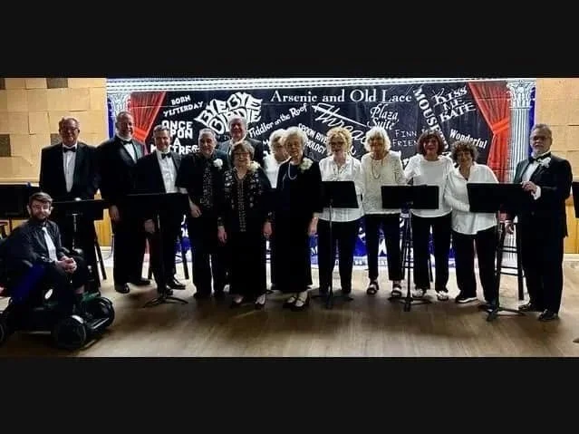 Group of 12 people, mostly women, standing on a stage holding music stands, with a man seated on the floor to the left, in front of a black backdrop with musical phrases and words. The people are dressed in formal and semi-formal attire, some in tuxedos and others in white shirts and black pants.