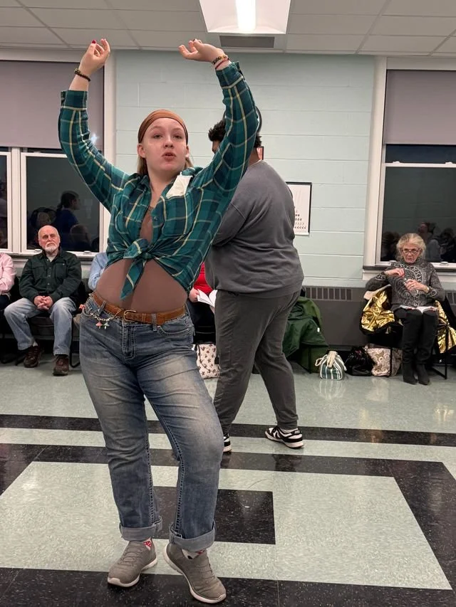 A young woman with red hair and a plaid shirt tied at the waist dancing in a room with an audience watching.