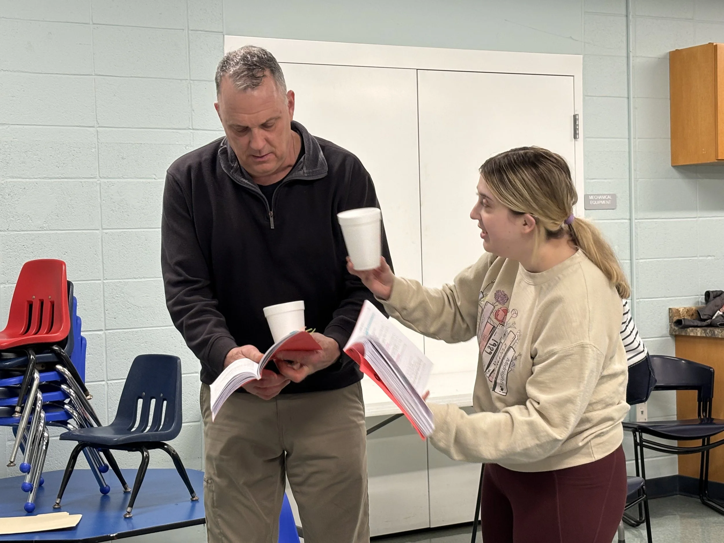 A man and a woman in a room with plastic chairs stacked along the wall, looking at documents together, with the woman handing a cup to the man.