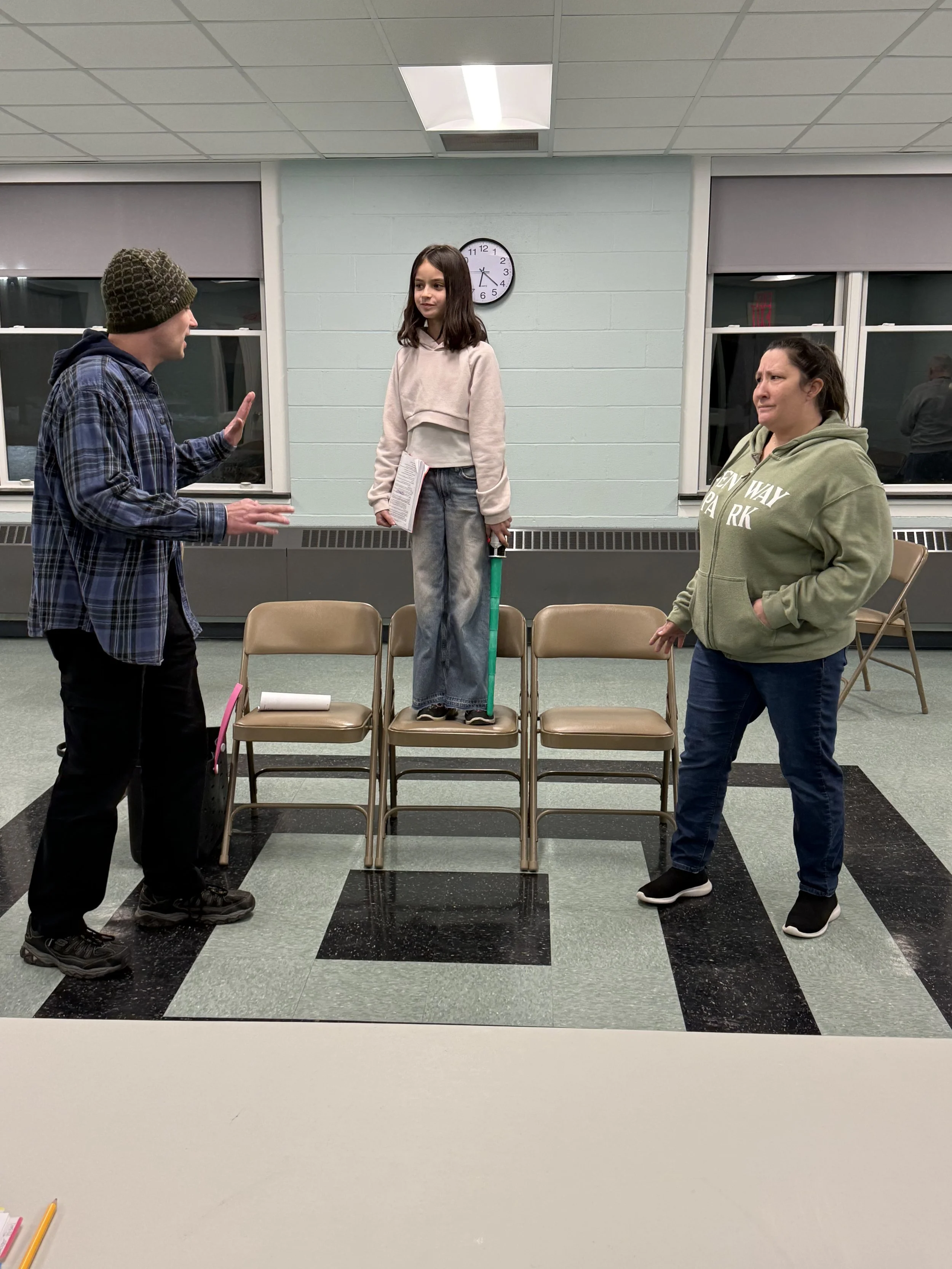 Three people in a room with chairs, two women and one girl standing on chairs. The girl is in the center, wearing a beige hoodie and jeans, holding a green stick and a book. The man on the left, wearing a knit hat and plaid jacket, is gesturing with his hand. The woman on the right, with a green hoodie, is listening with a serious expression. A clock on the wall shows approximately 4:11.
