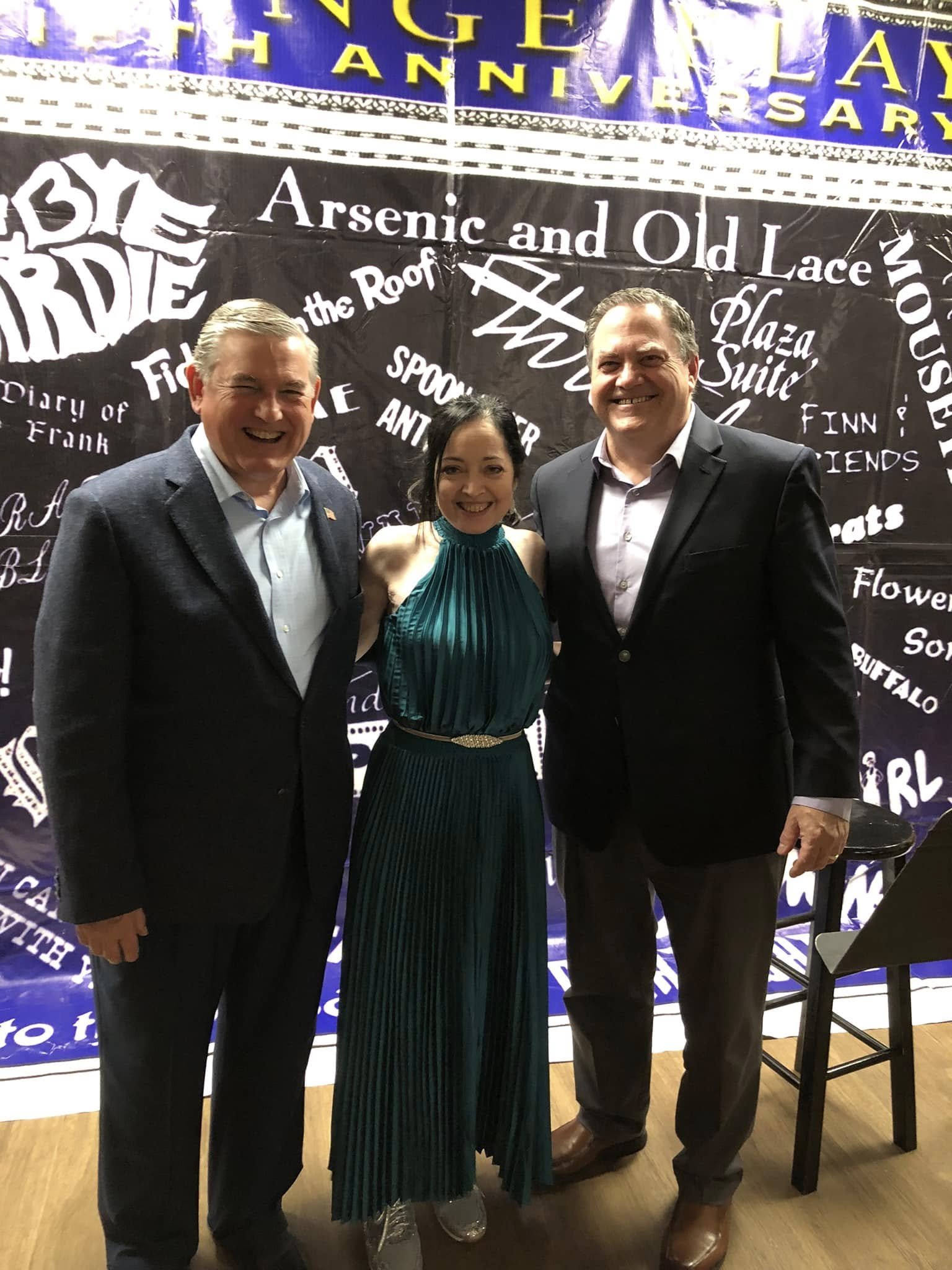 Three smiling people standing in front of a black banner with white text, at a celebration event.