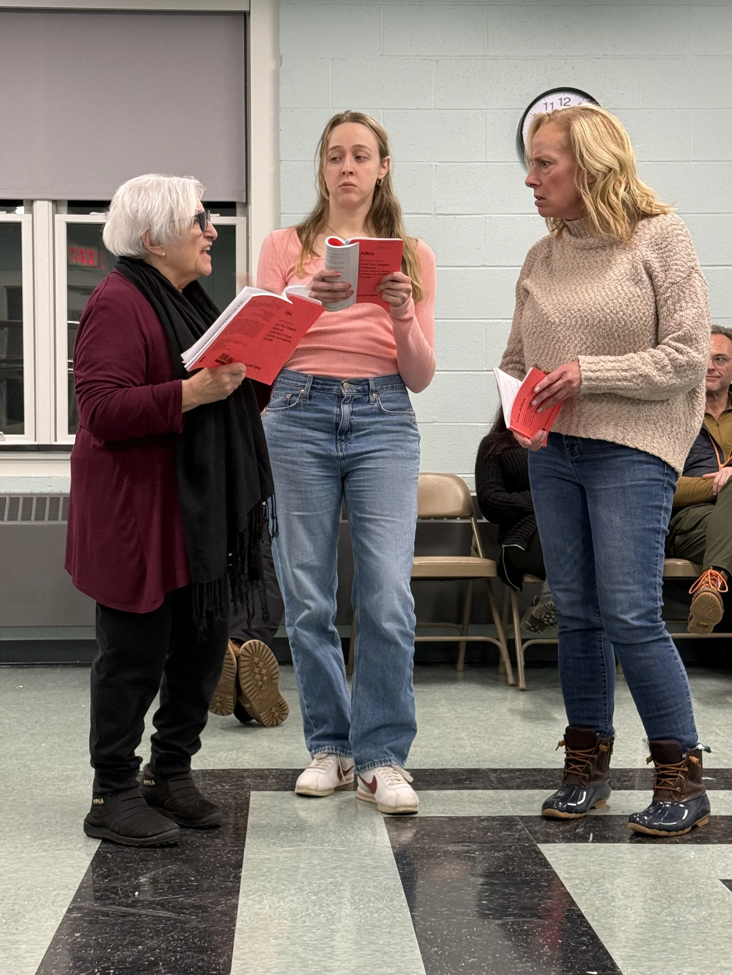 Three women standing in an indoor setting, engaged in conversation, holding red-books, with a few seated individuals in the background, and a clock on the wall.