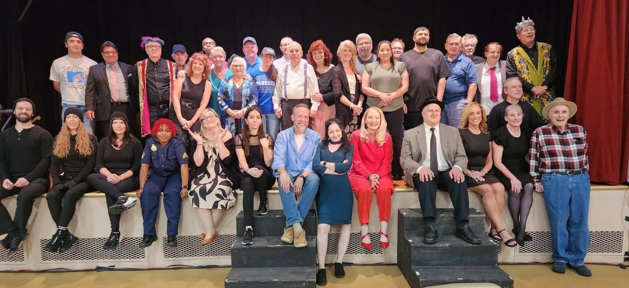Group of people on a stage, some sitting and some standing, posing for a photo after a performance or event. Many are smiling and dressed in casual or costume attire, with a red curtain in the background.