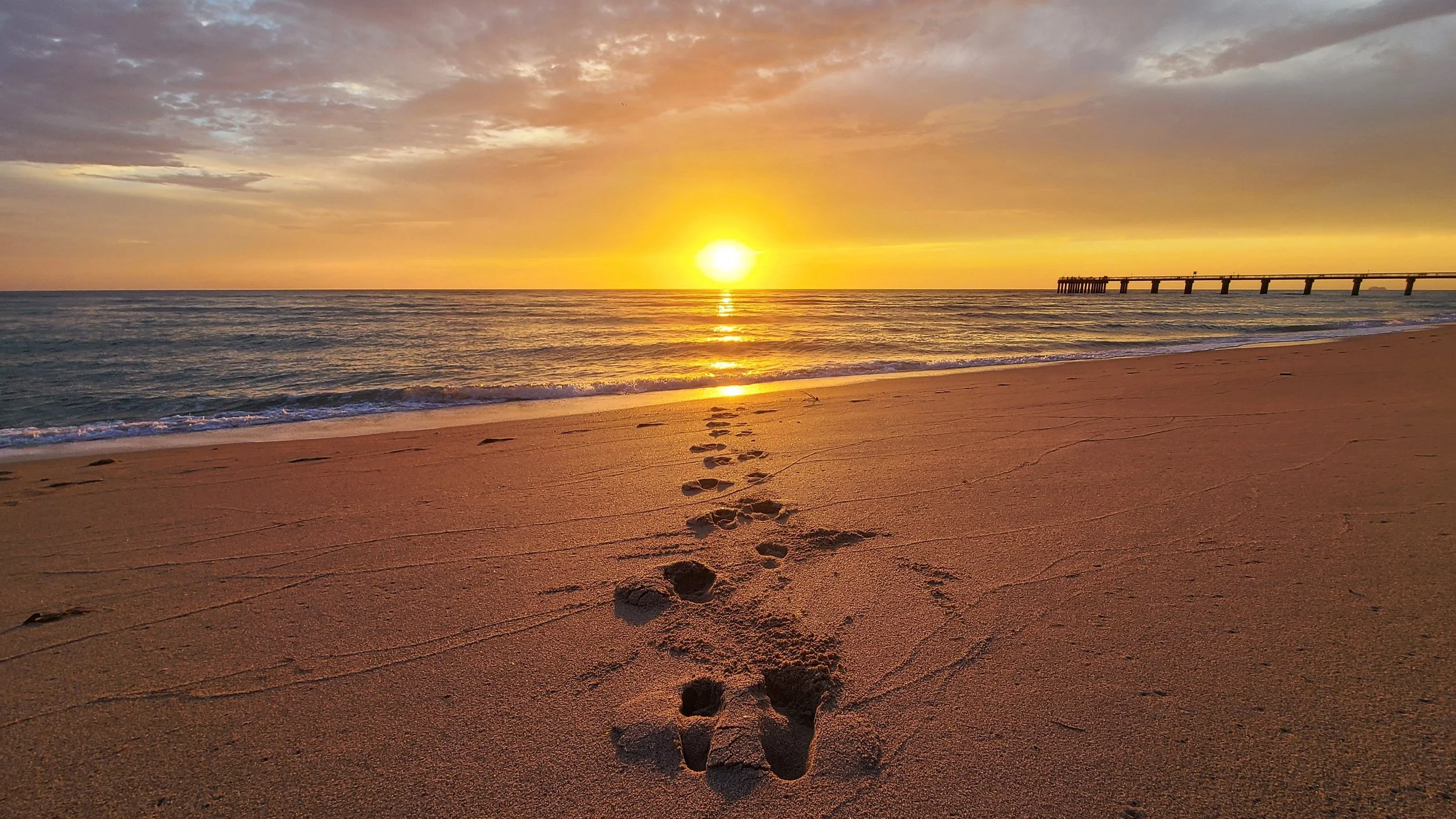 Sunrise from the beach with the Newport Fishing Pier on the right. Footsteps are leading up to the ocean.