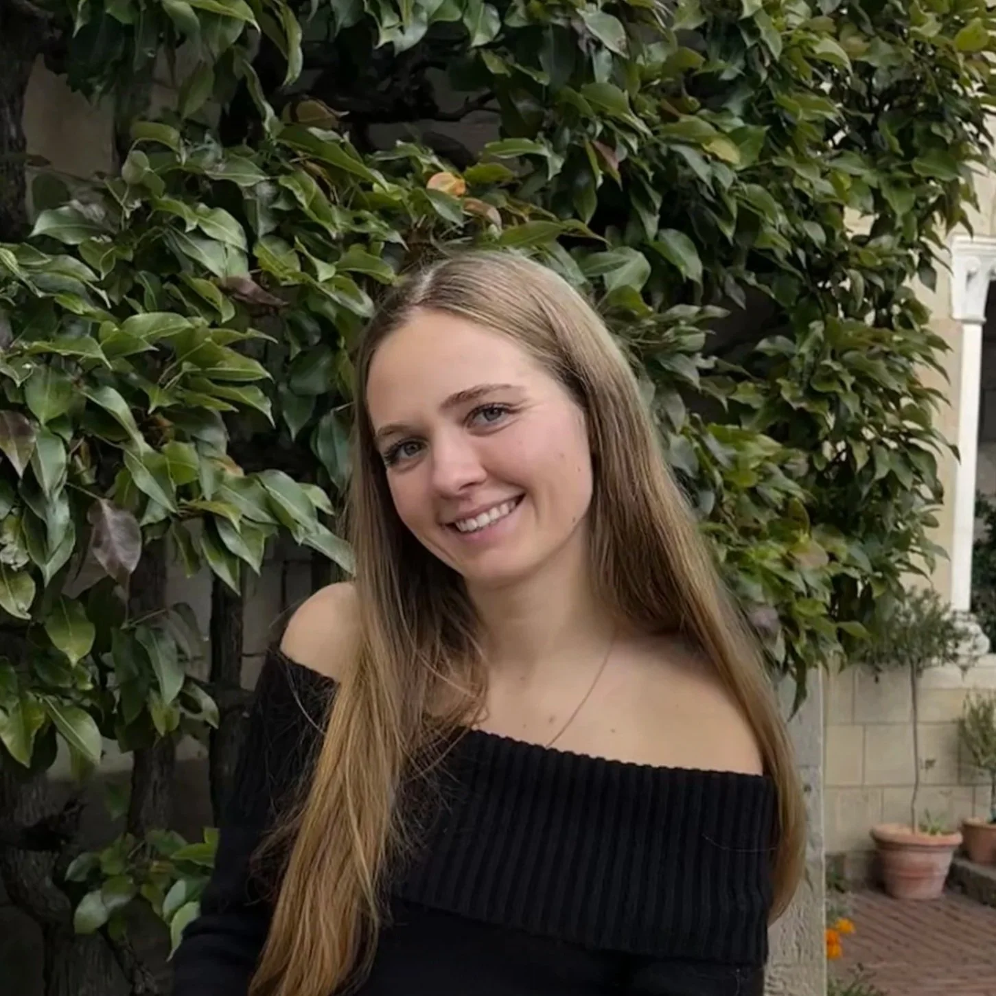 A young woman with long blonde hair and blue eyes smiling in front of a hedge of green leaves, wearing a black off-the-shoulder top.