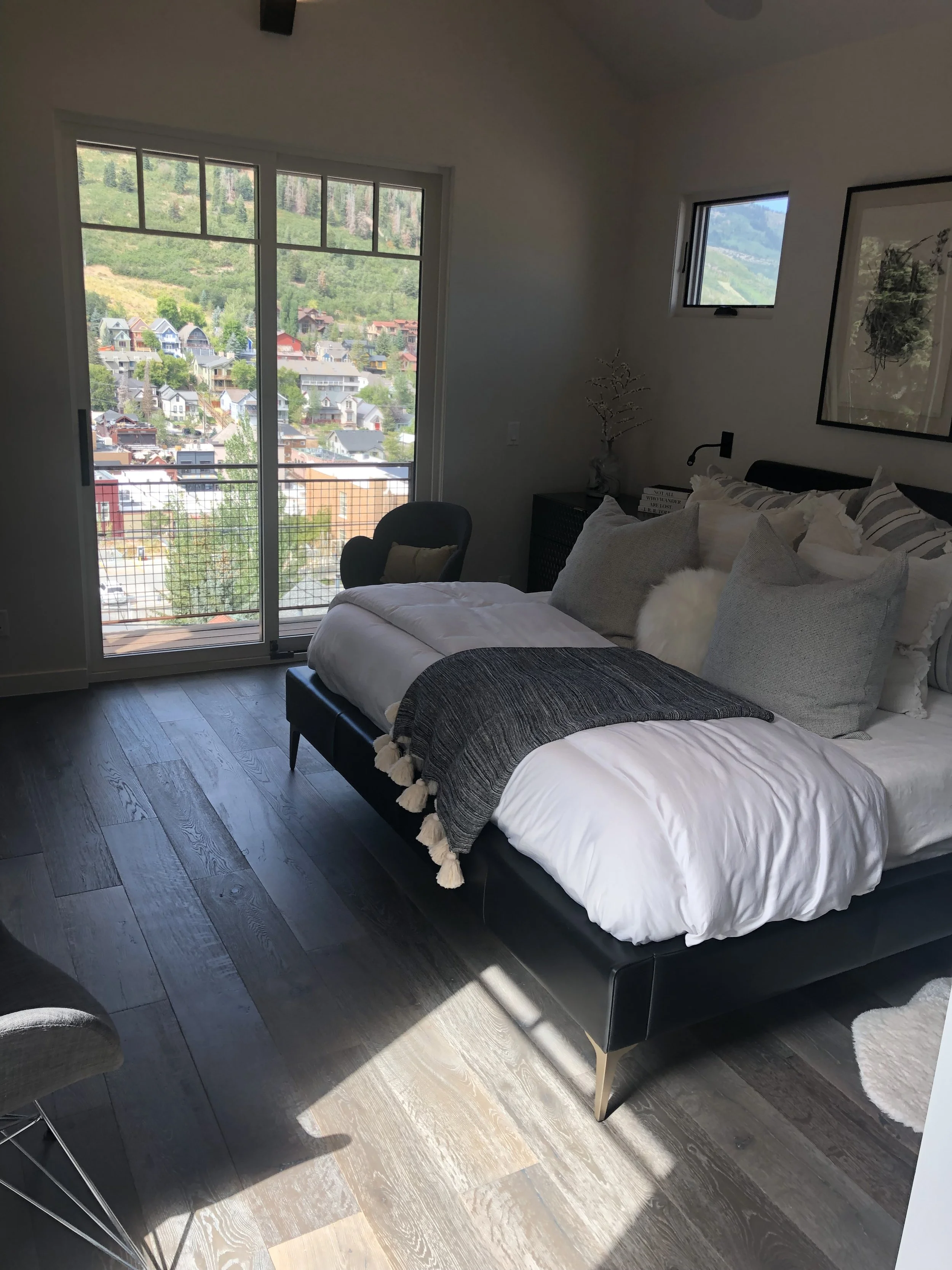 Living room with a large window showing a view of a hillside with houses, a bed with white and gray pillows and blanket, dark wooden flooring, and a small side table with a plant and books.