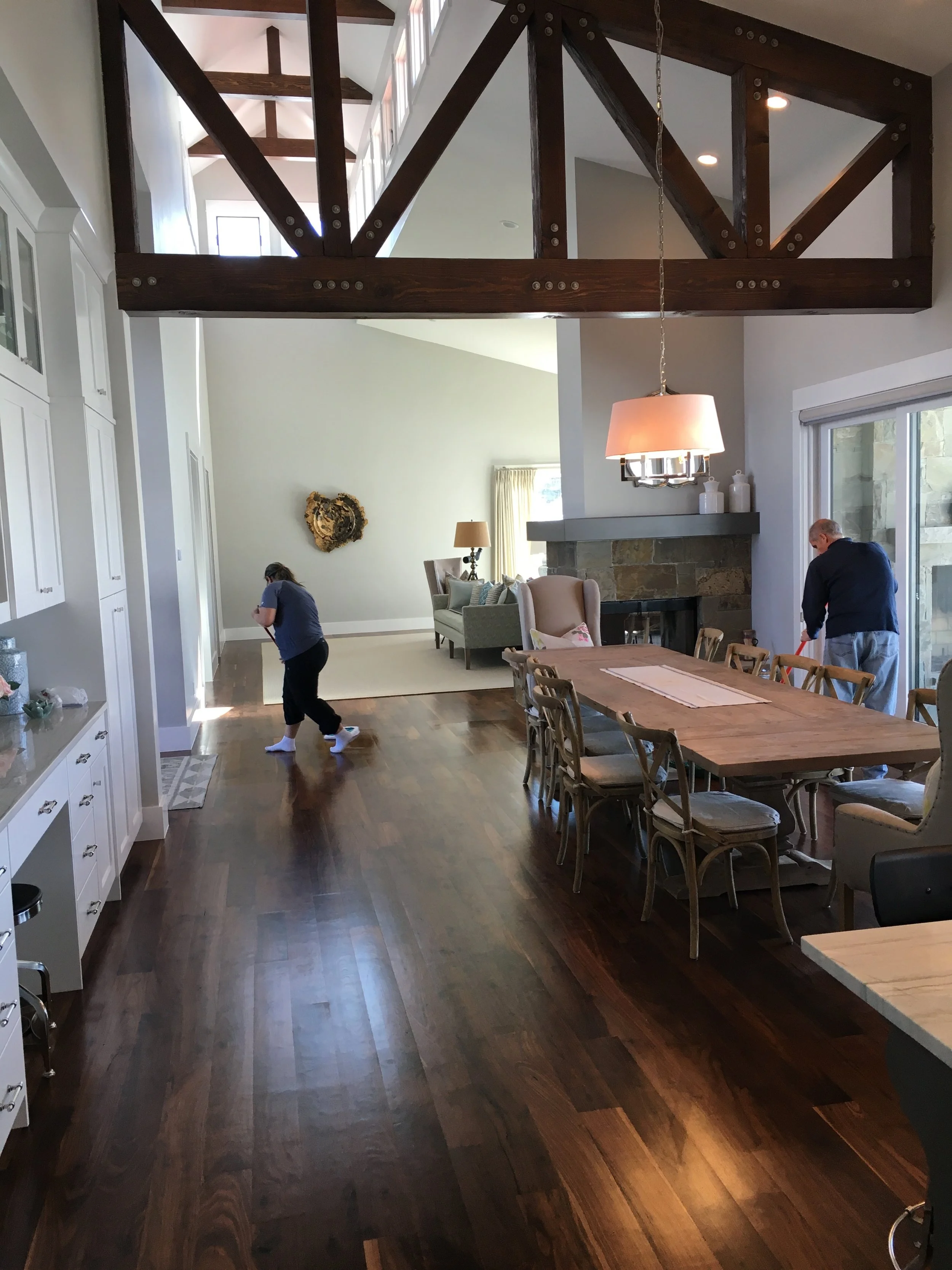 A spacious living room with a high ceiling and wooden beams. There is a dining table with chairs, a fireplace, large windows letting in natural light, and people cleaning the floor.
