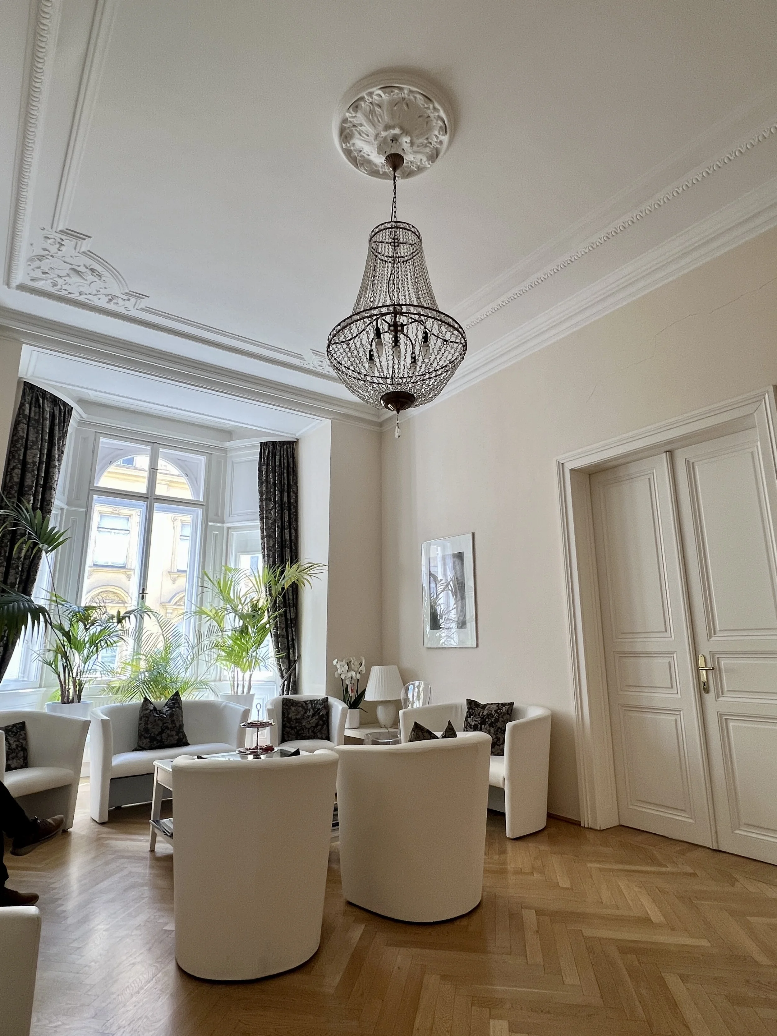 Ein eleganter Raum mit weißen Sesseln, schwarzen Kissen, einem kleinen Tisch, einem großen Fenster mit schwarzen Vorhängen, einem Kronleuchter und einer weißen Wand mit einem dekorativen Rahmen. Pflanzen stehen an der Fensterbank.