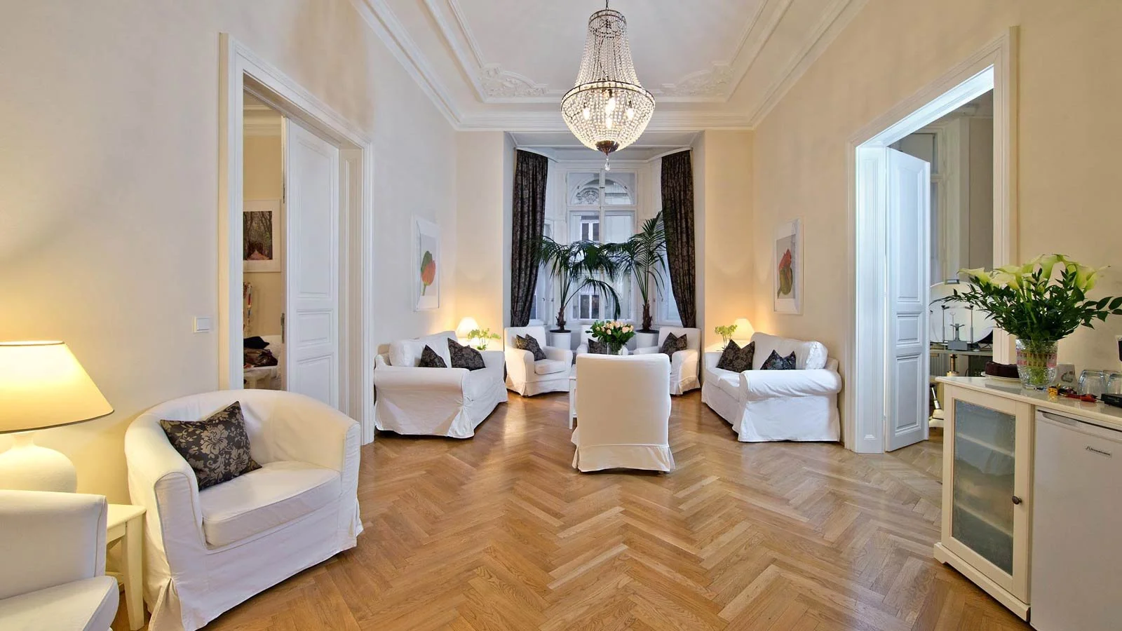 Elegantes Wohnzimmer mit weißen Sofas, dunklen Kissen, großer Deckenleuchte, Parkettboden, hohen Fenstern mit Vorhängen und Pflanzen.