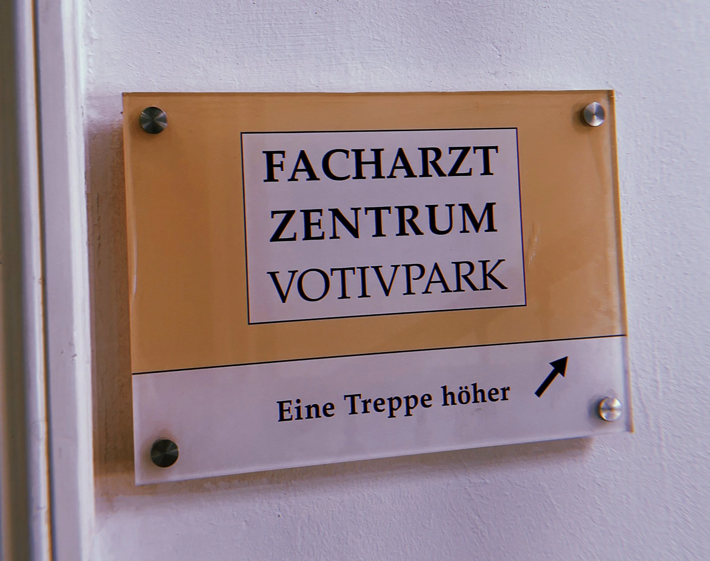 Hinweisschild an der Wand mit schwarzer Umrandung, orangefarbenem Hintergrund und schwarzer Schrift, auf dem steht 'Facharzt Zentrum' und darunter weitere Hinweise.
