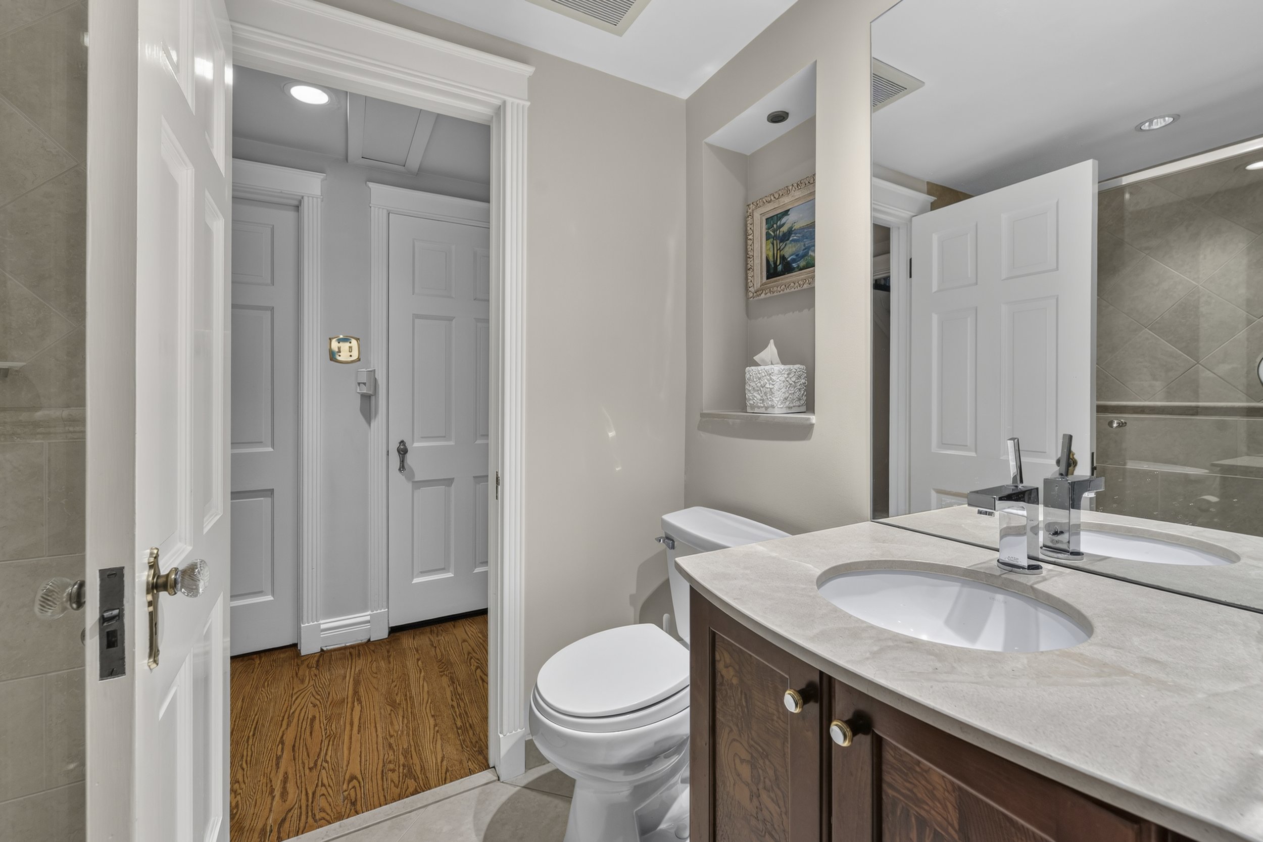 A bathroom with a double sink vanity, toilet, and a mirror. The walls are painted white, and there is a framed painting on the wall. The bathroom has wooden flooring and a shower visible in the mirror reflection.