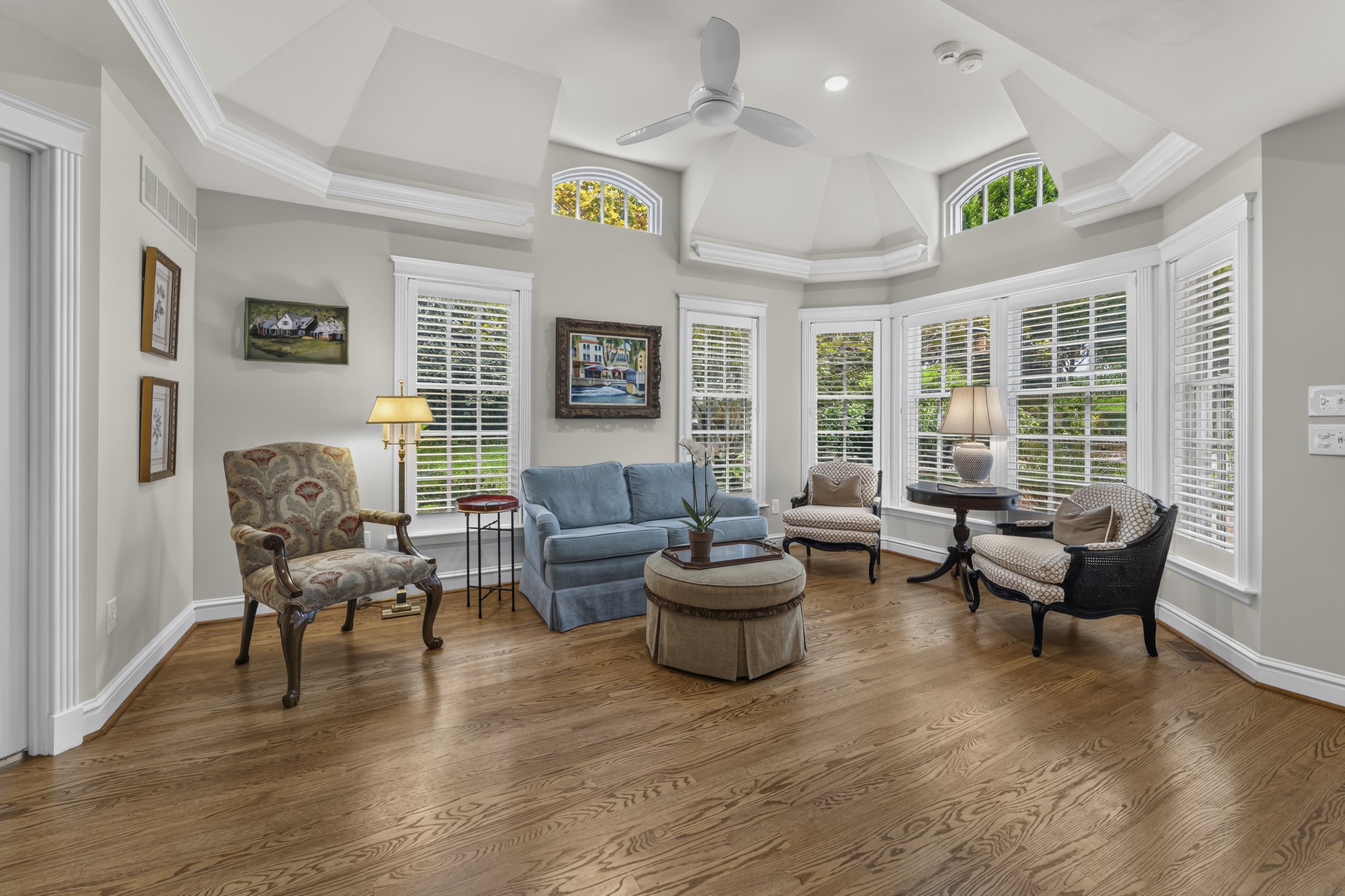 Living room with hardwood floors, various chairs, a blue couch, side tables, and artwork on the walls. Large windows with white blinds let in natural light.