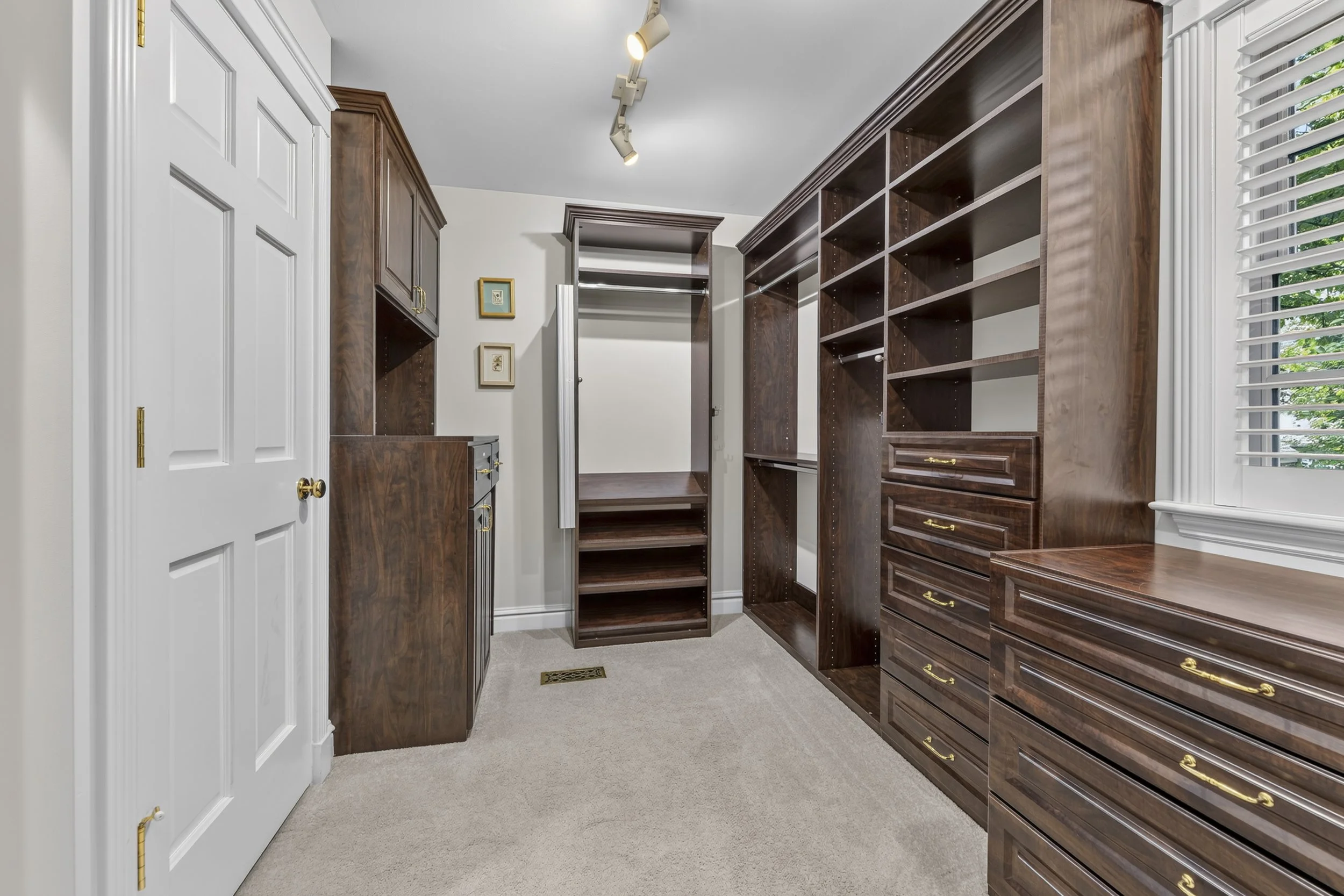 Empty walk-in closet with wooden storage shelves, drawers, and a white door with gold handles. A window with white blinds and a small framed picture on the wall are visible.
