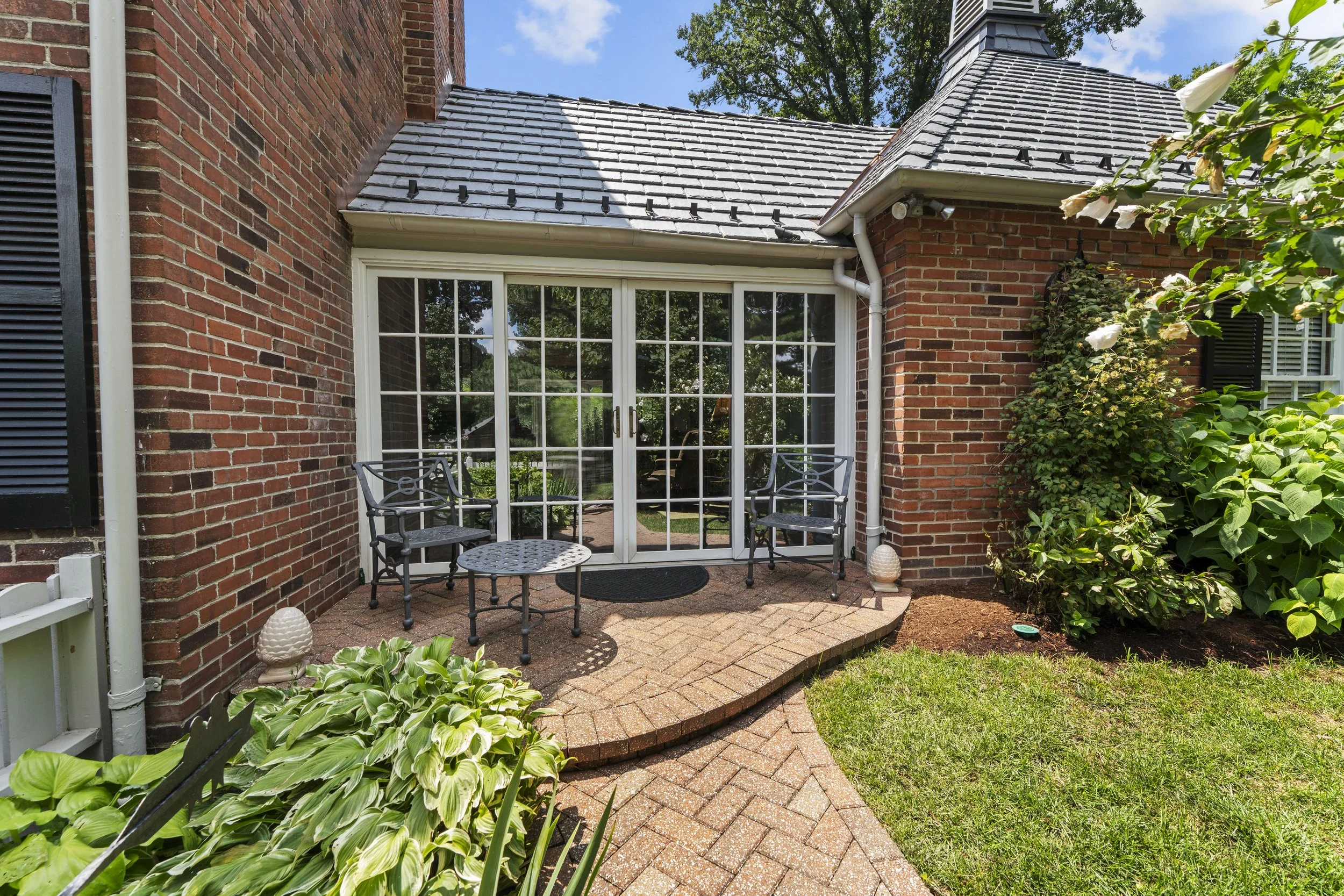 Backyard patio with brick flooring, black chairs, and a sliding glass door leading into a house with brick walls and a shingled roof. Surrounding greenery includes bushes, a tree, and potted plants.