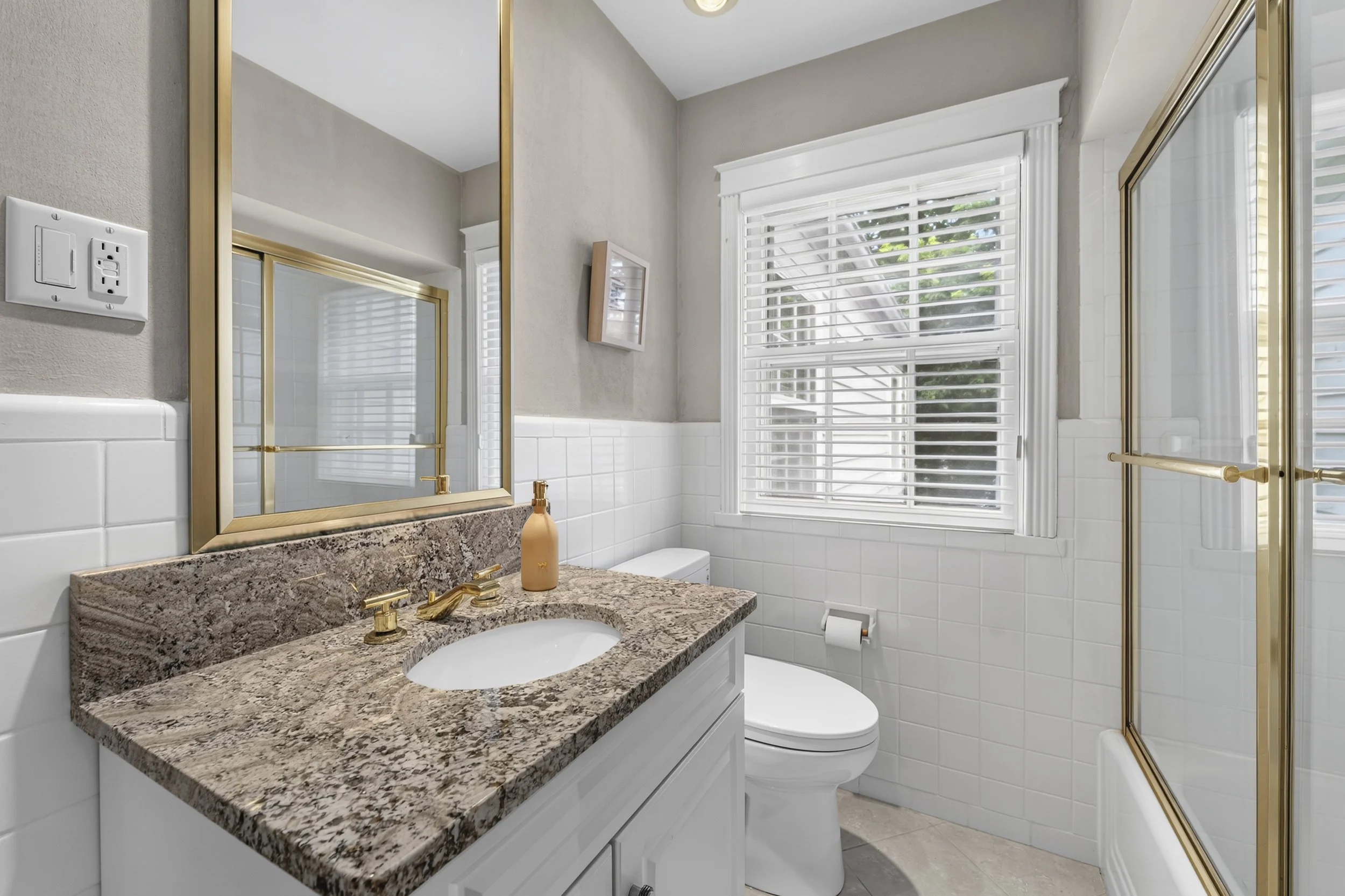 A bathroom featuring a granite countertop with a built-in sink, gold faucet, and soap dispenser, with a large mirror above. The room has white tiled walls, a white toilet, and a window with white blinds, allowing natural light to enter. There are gol