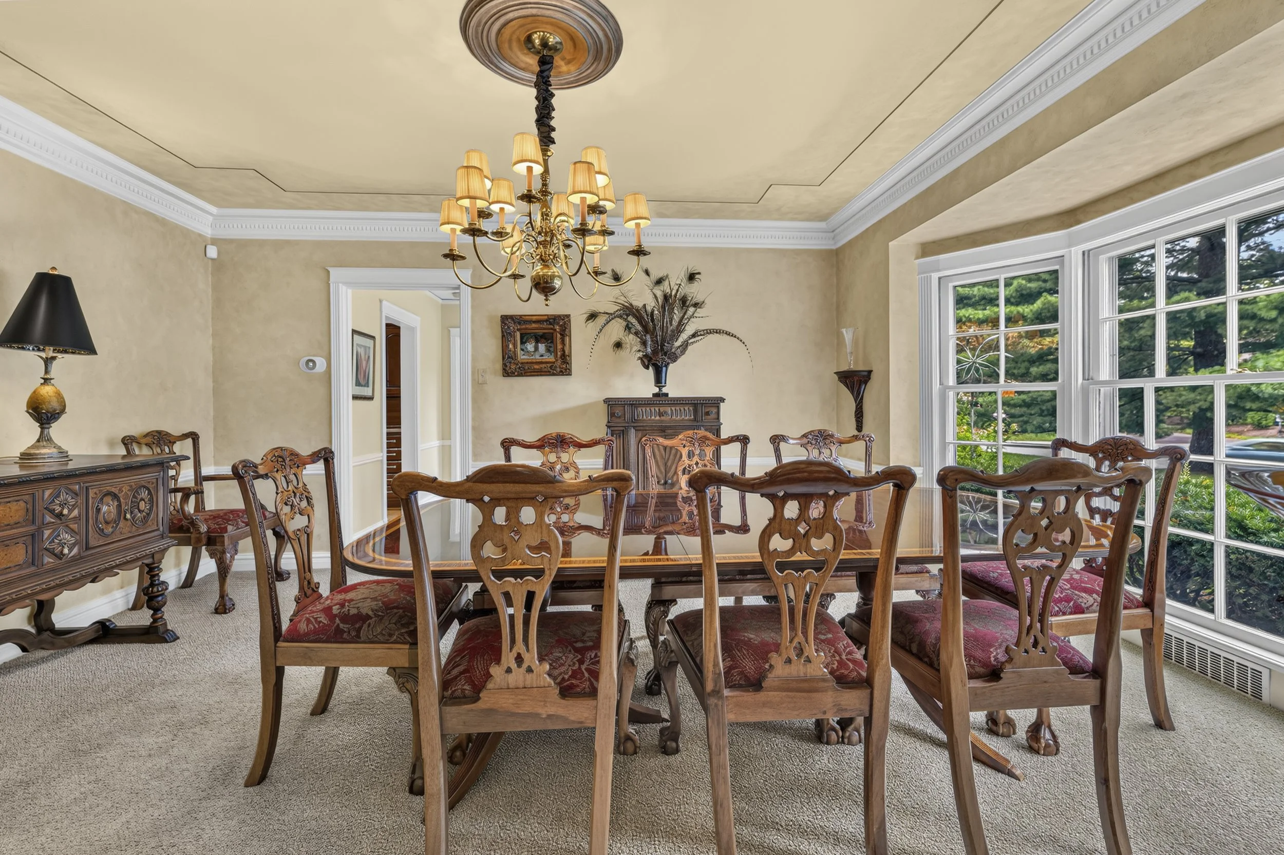 A traditional dining room with a large wooden dining table surrounded by carved wooden chairs with red cushioned seats. The room features a chandelier hanging from a wood-paneled ceiling, large bay windows with a view of greenery outside, and a sideb