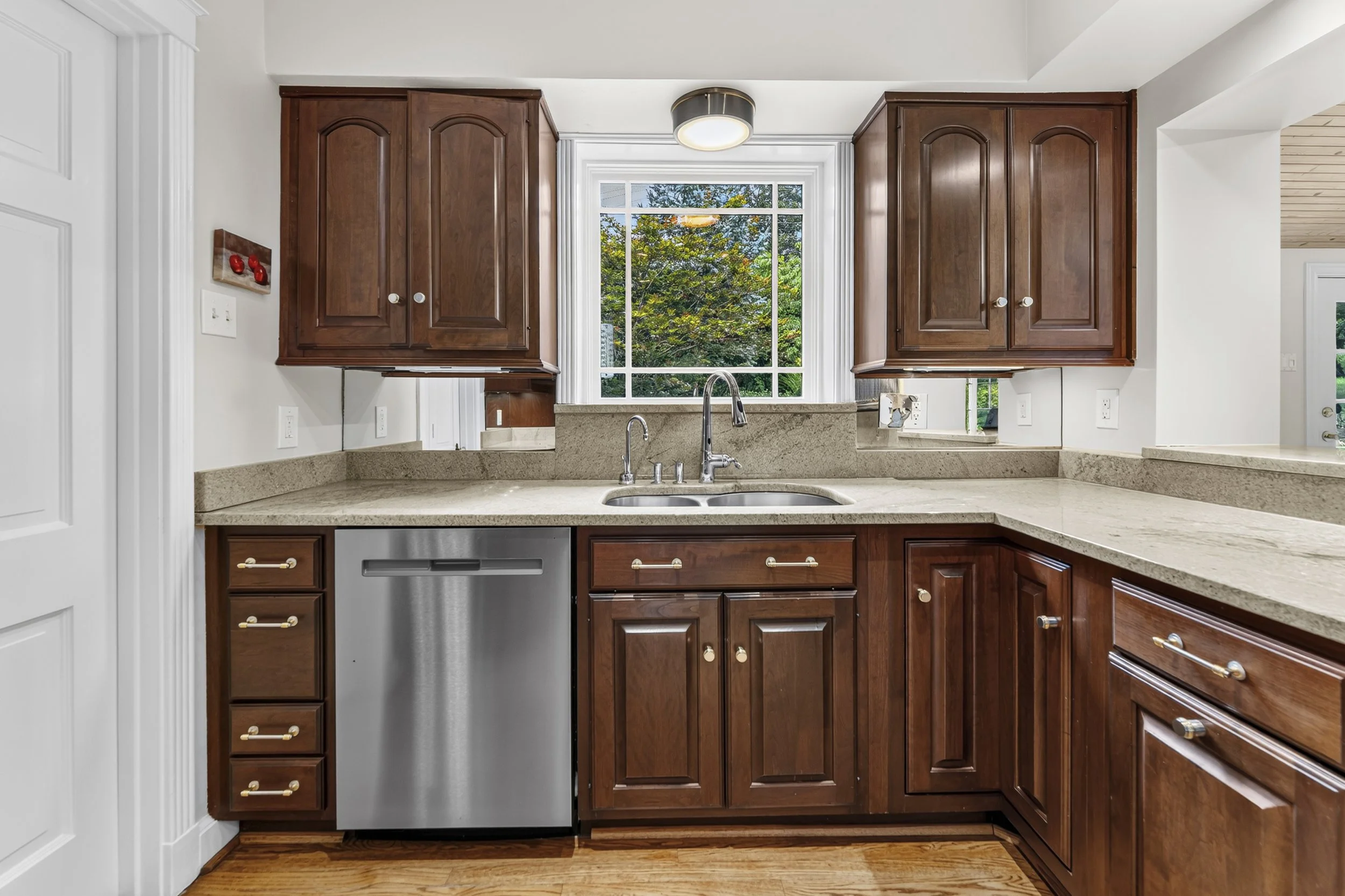 Kitchen with dark wood cabinets, granite countertops, a window showing green trees, and a stainless steel dishwasher.