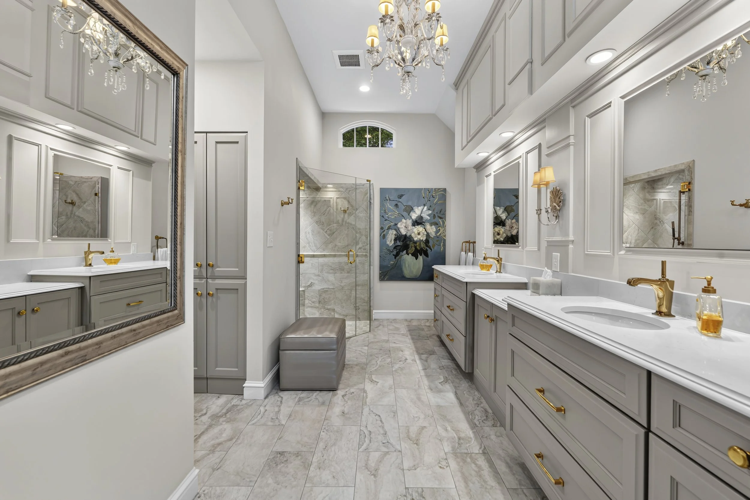 A modern, elegant bathroom with white walls, marble flooring, and gray cabinetry with gold hardware. The bathroom features double sinks, a large mirror, and a chandelier hanging from the ceiling. There is a framed mirror on the left wall, a floral pa