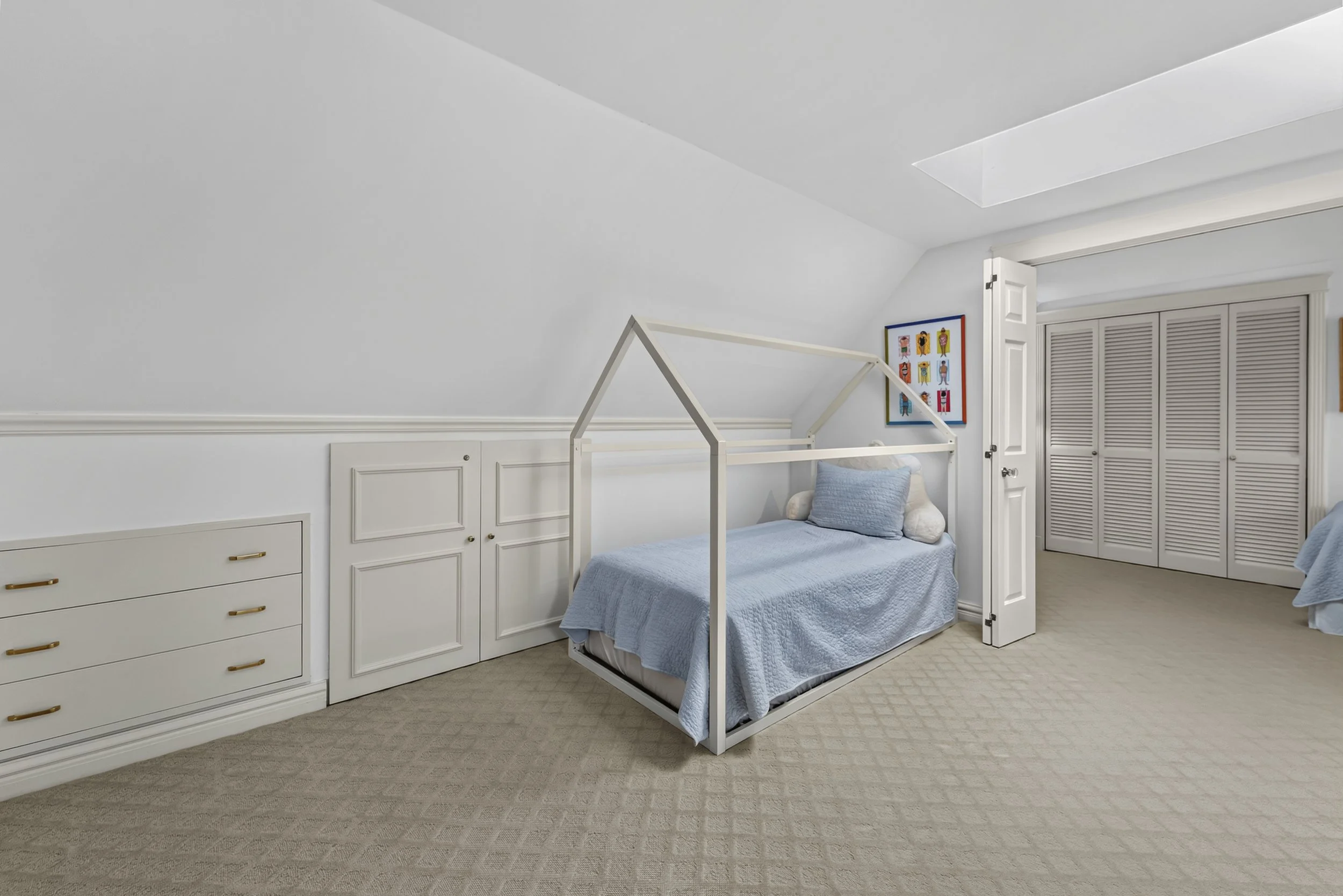 A bedroom with a white house-shaped bed frame, light blue bedding, and pillows. Art on the wall features colorful characters. White trim, closet doors, and a carpeted floor.