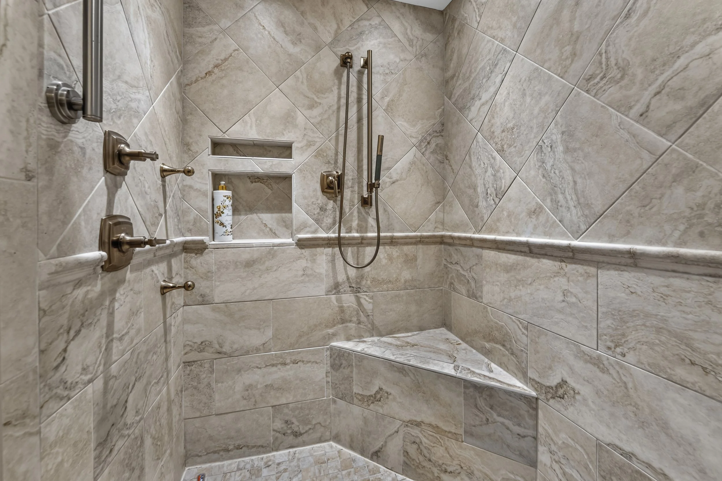 A tiled shower with beige and brown marble patterns, featuring a built-in corner shelf with shower products, two showerheads, and a grab bar.