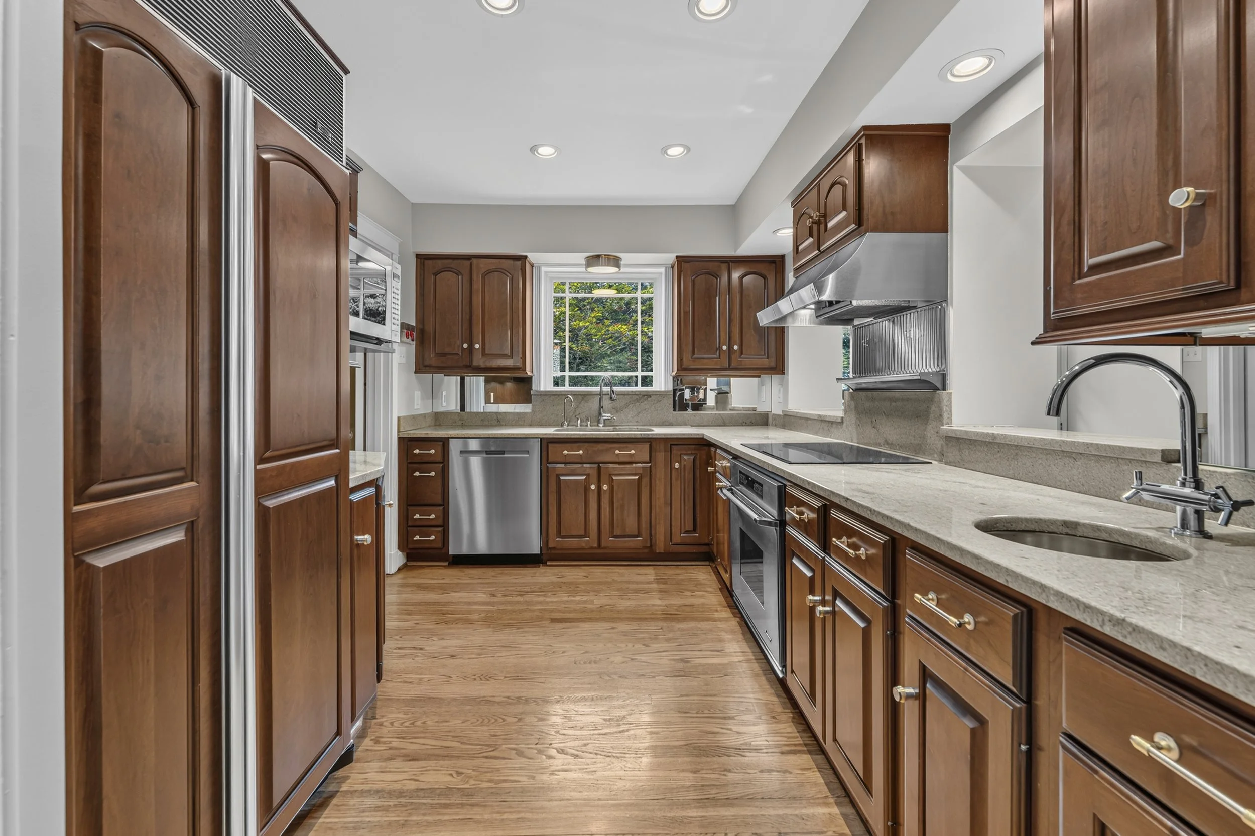 View of a kitchen with wooden cabinets, beige countertops, a stainless steel dishwasher, and a window with greenery outside.