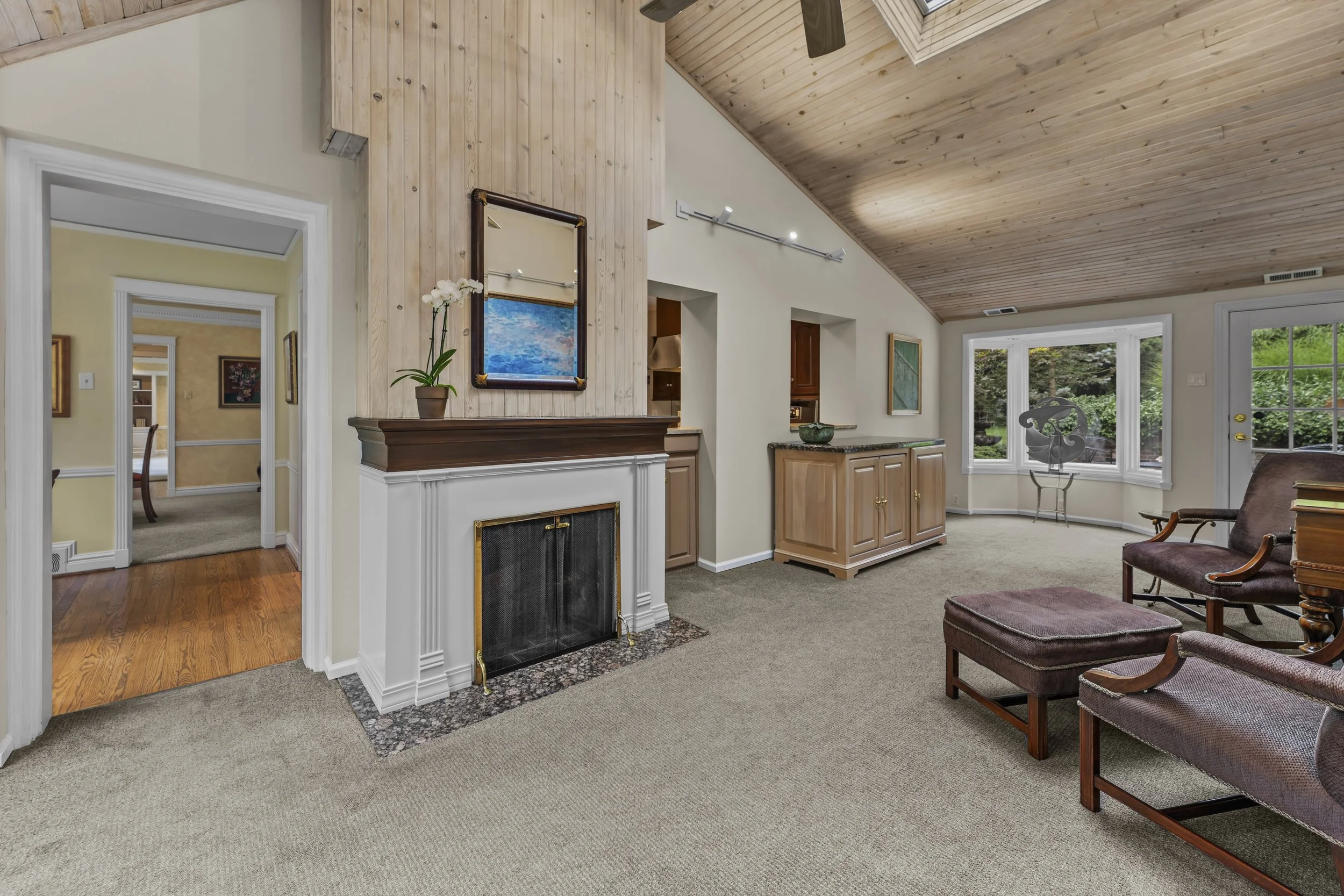 Living room with beige carpet, wood-paneled vaulted ceiling, and large window showing a green outdoor yard. There is a fireplace with a wooden mantle, a potted orchid, and artwork on the walls. A brown cushioned chair with wooden arms is partially vi