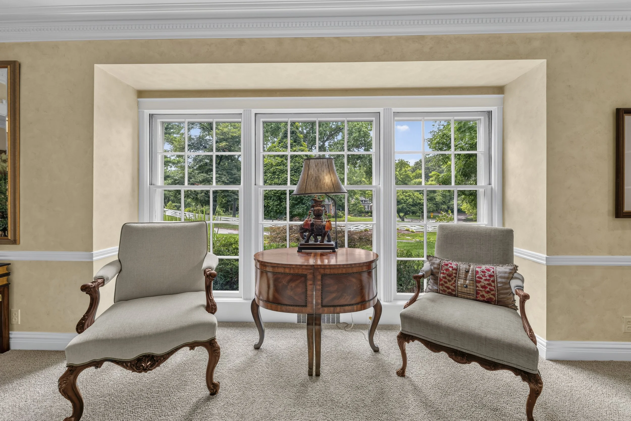 Living room with two upholstered armchairs with carved wooden legs facing a round wooden table with a decorative table lamp, large double-pane window with views of greenery, and framed artwork on the walls.