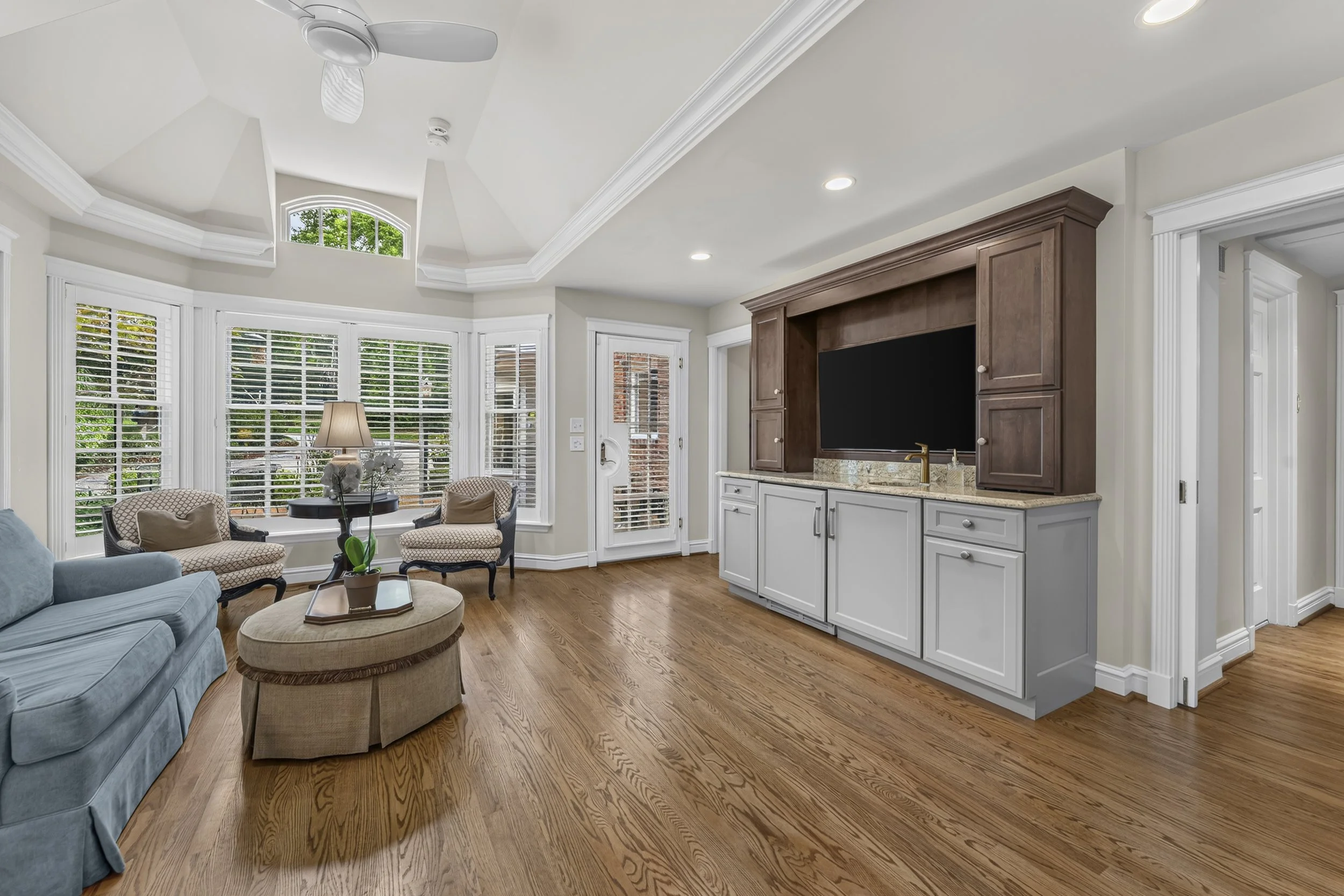 Living room with large front windows, beige and blue furniture, wooden floors, white cabinetry with a television, ceiling fan, and a view of trees outside.