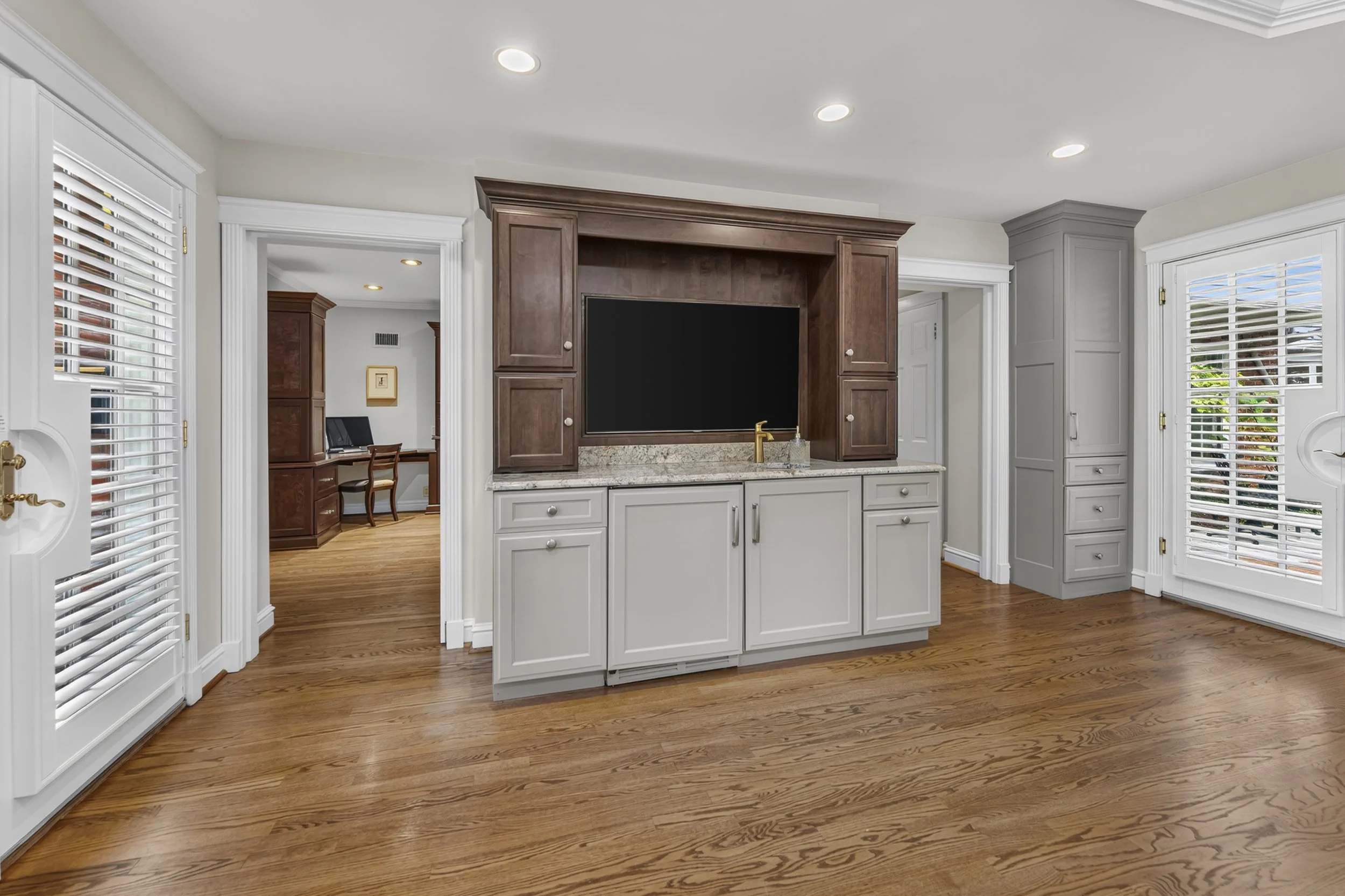 Living room with hardwood floors, white and brown cabinets, a television, and large windows with white blinds.