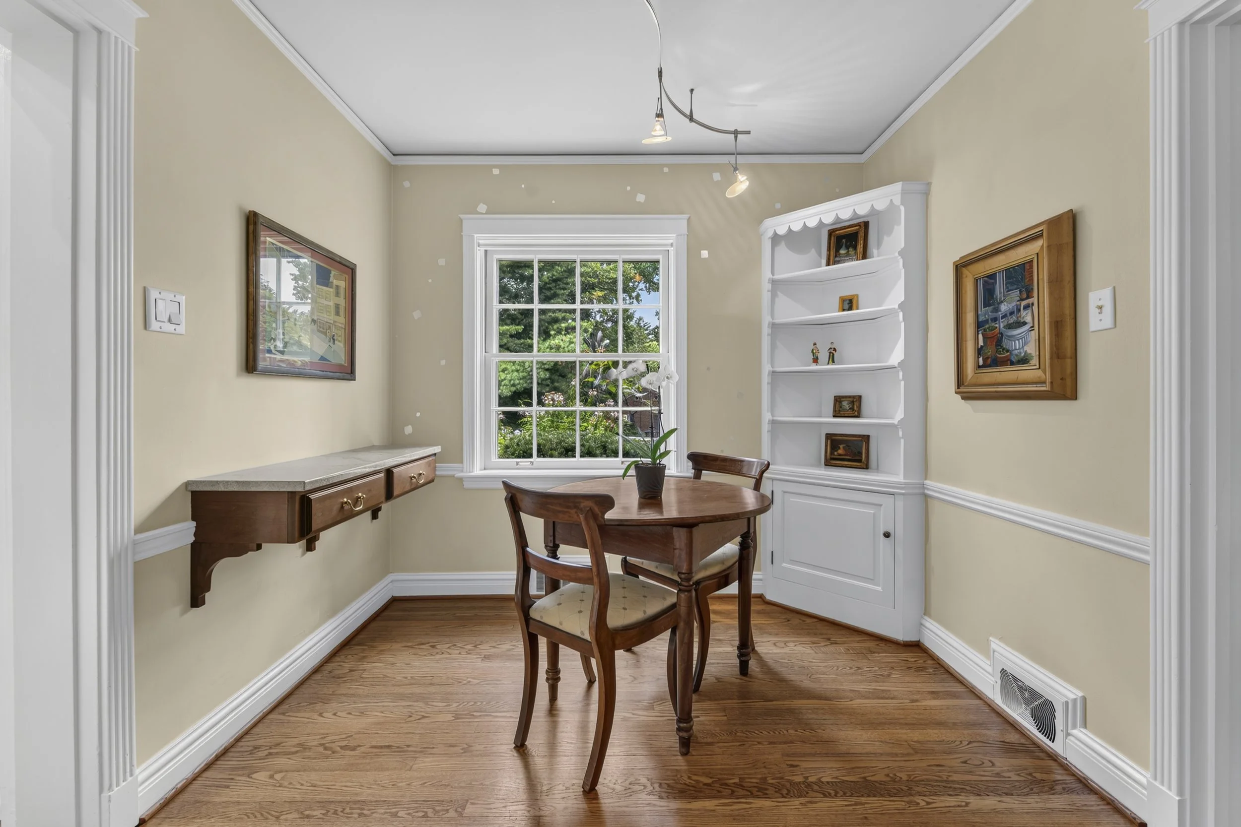 Interior of a dining room with a round wooden table and three chairs, a window with a green view, decorative white shelf with small paintings, framed artwork on beige walls, hardwood floors, and track lighting on the ceiling.