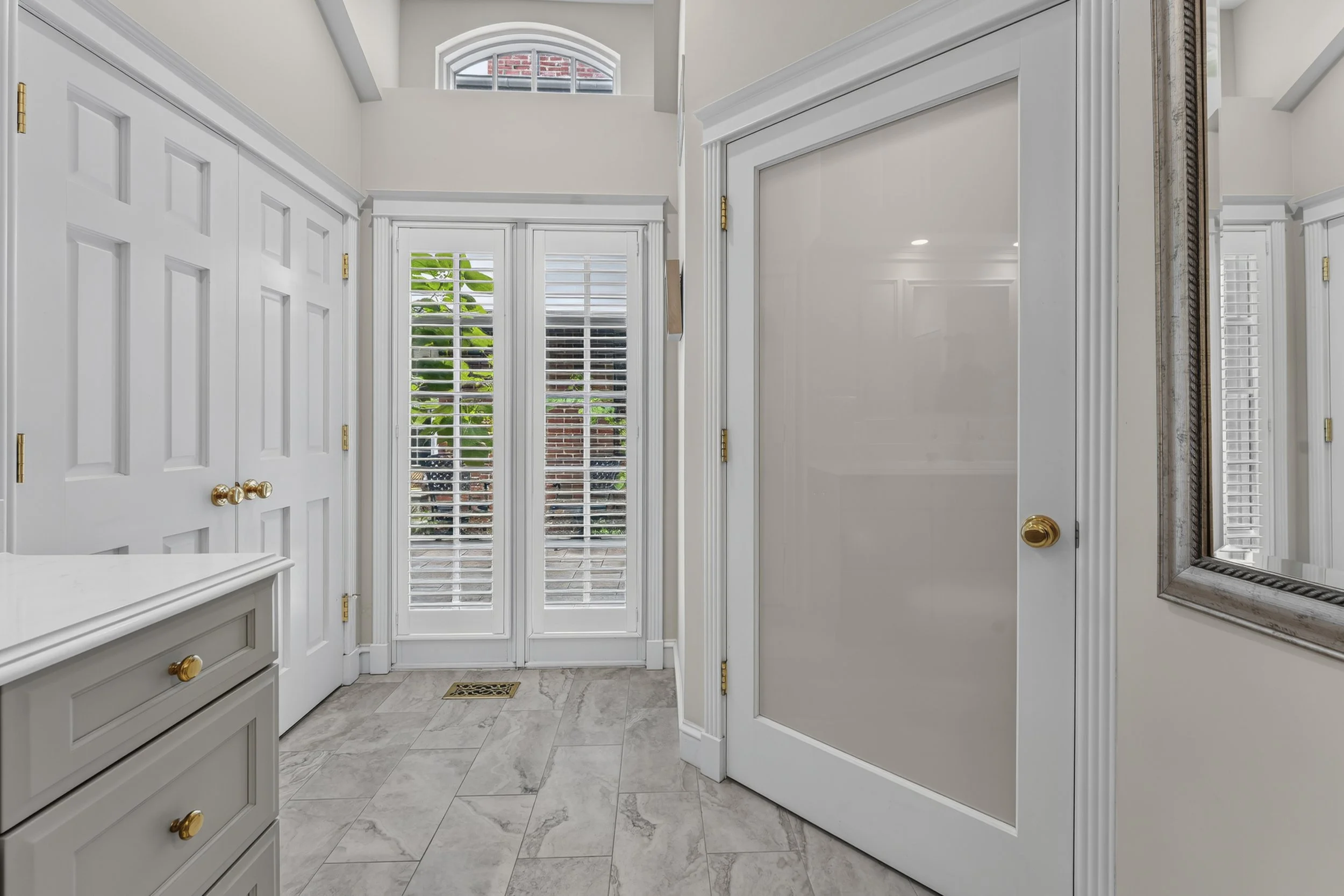 A bright foyer with white walls, white doors with gold handles, a silver framed mirror, marble-tile flooring, and a glass door with white plantation shutters leading outside, where plants are visible.