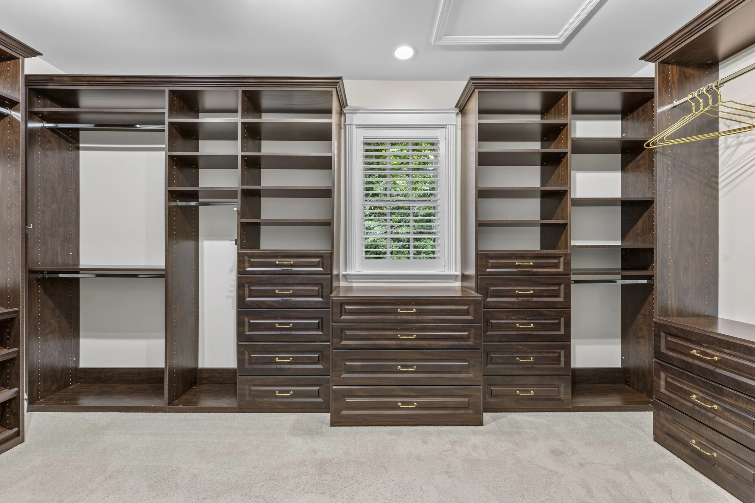 Empty walk-in closet with built-in wooden shelves and drawers, a window with white blinds, and beige carpet.