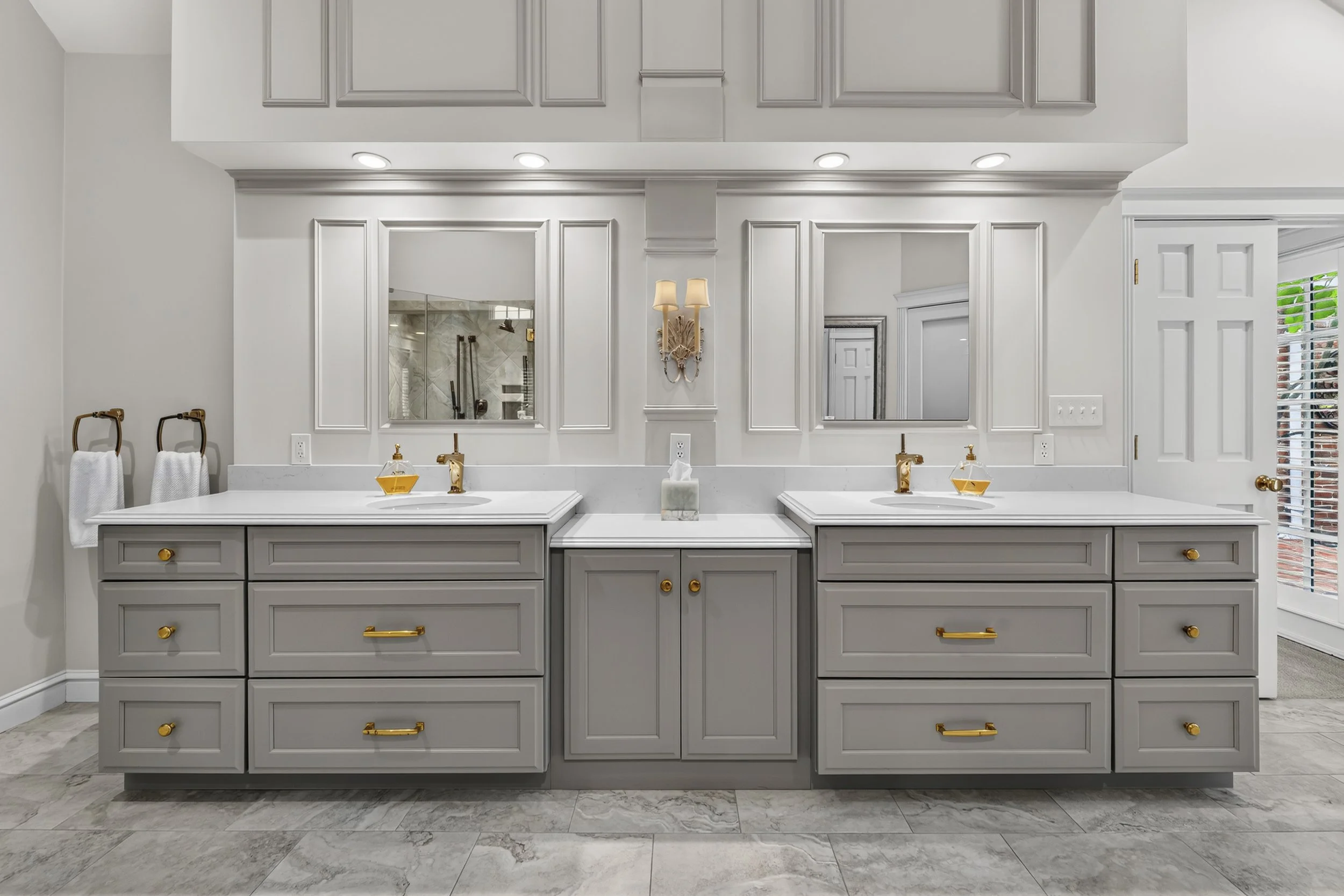 A modern double vanity bathroom with gray cabinets, gold hardware, and a marble countertop. There are two sinks with gold faucets, mirrors, and wall-mounted lights. Towels hang on gold towel bars, and a tissue box is on the counter. The bathroom has 