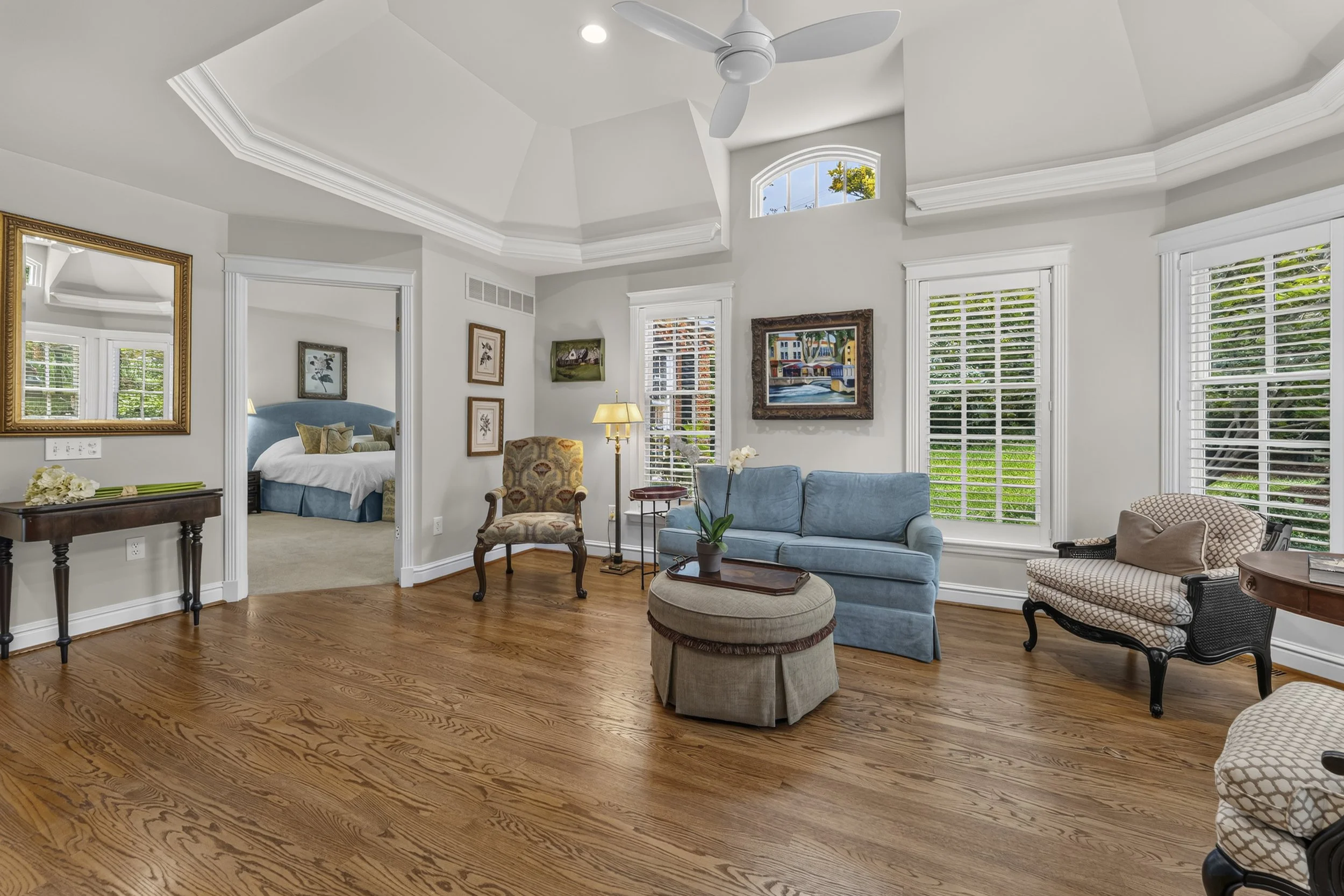 Bright living room with wooden floors, large windows with white blinds, a blue sofa, patterned armchairs, paintings on the walls, a wooden coffee table, and a ceiling fan.