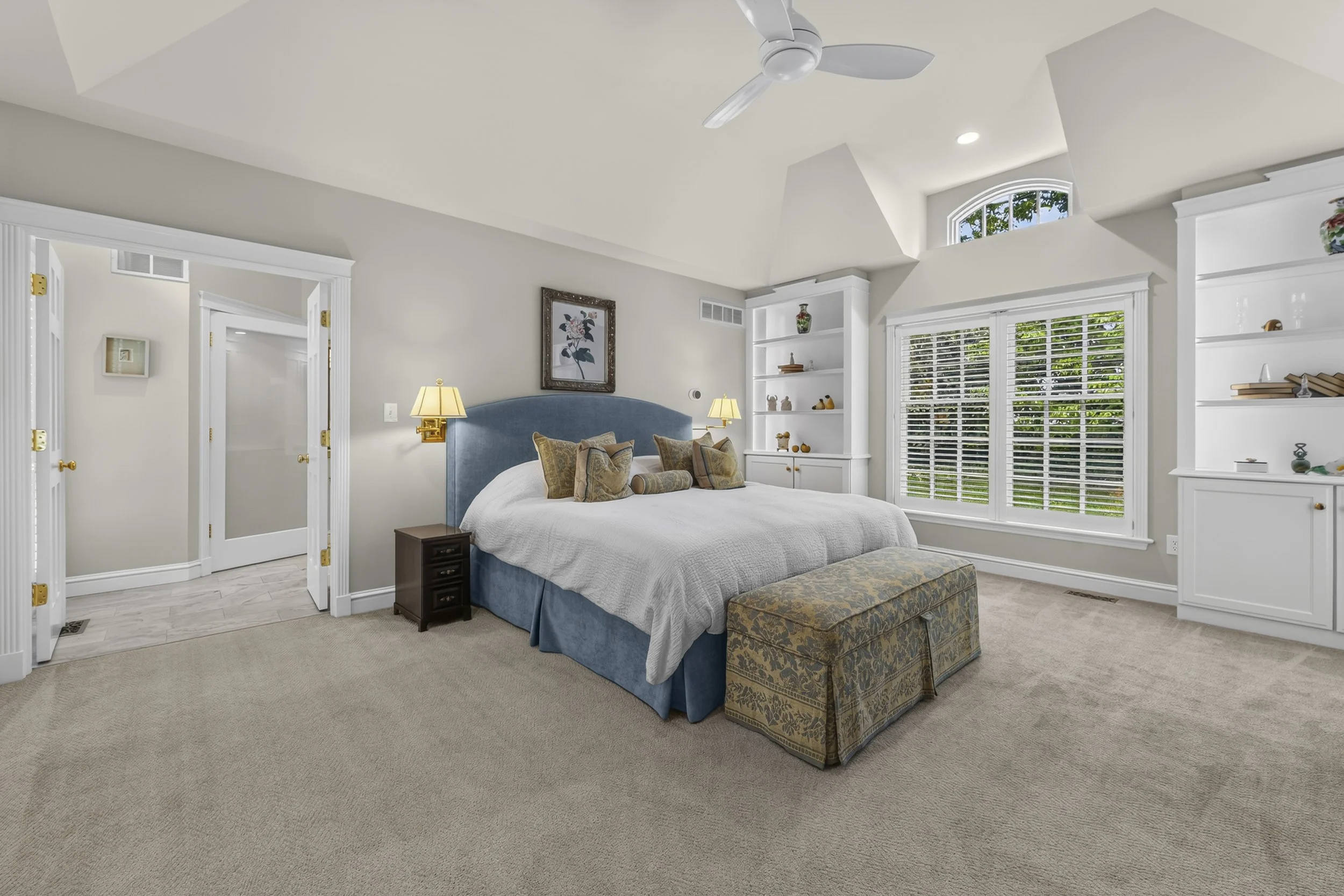 A bright bedroom with a white ceiling, sloped roof, and a ceiling fan. There is a large window with white blinds and a view of green foliage outside. The bed has a blue upholstered headboard, white bedding, patterned pillows, and a padded bench at th