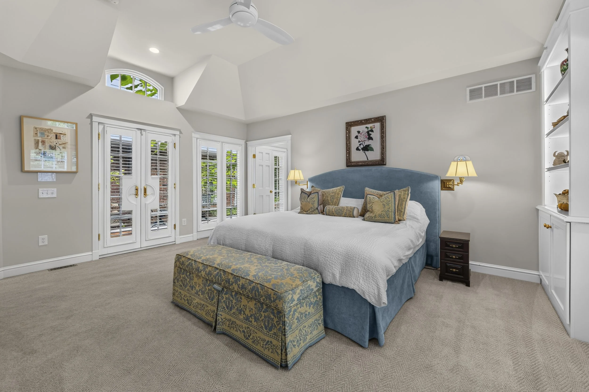 A bedroom with a large bed, blue upholstered headboard, and decorative pillows. There is an upholstered bench at the foot of the bed, white walls, and built-in shelving. A ceiling fan and two wall-mounted lamps accompany a window with white shutters 