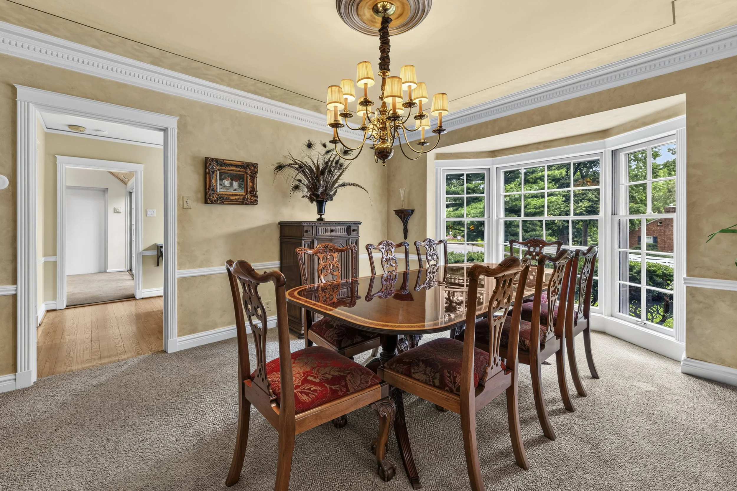 A dining room featuring a wooden table with ornate wooden chairs upholstered with red patterned fabric, a large brass chandelier hanging from the ceiling, and a large bay window showing green trees outside. There's a sideboard with a decorative vase 