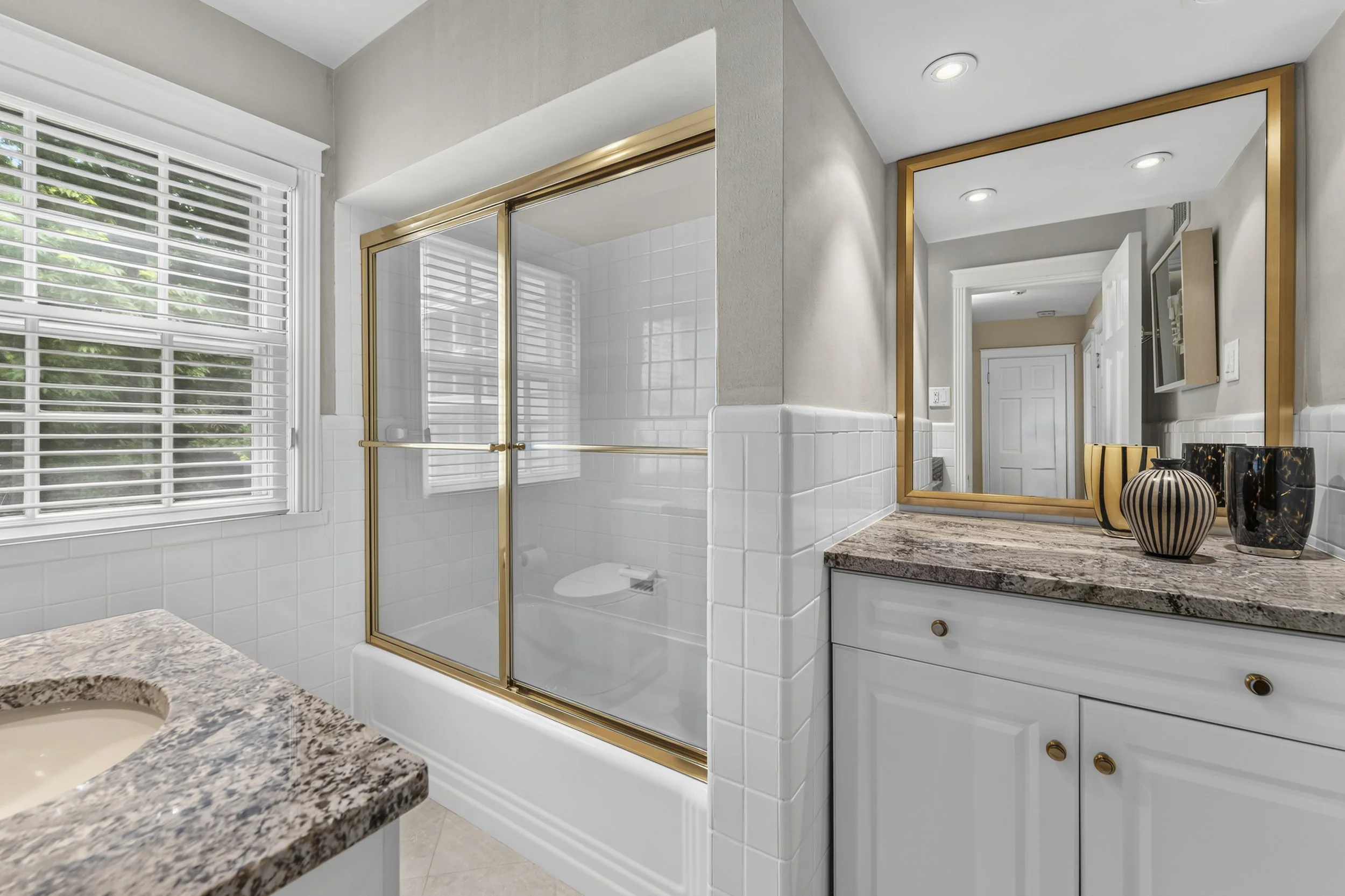 Bright bathroom with large window with white blinds, porcelain sink with granite countertop, and gold-framed mirror with decorative vases on the countertop.