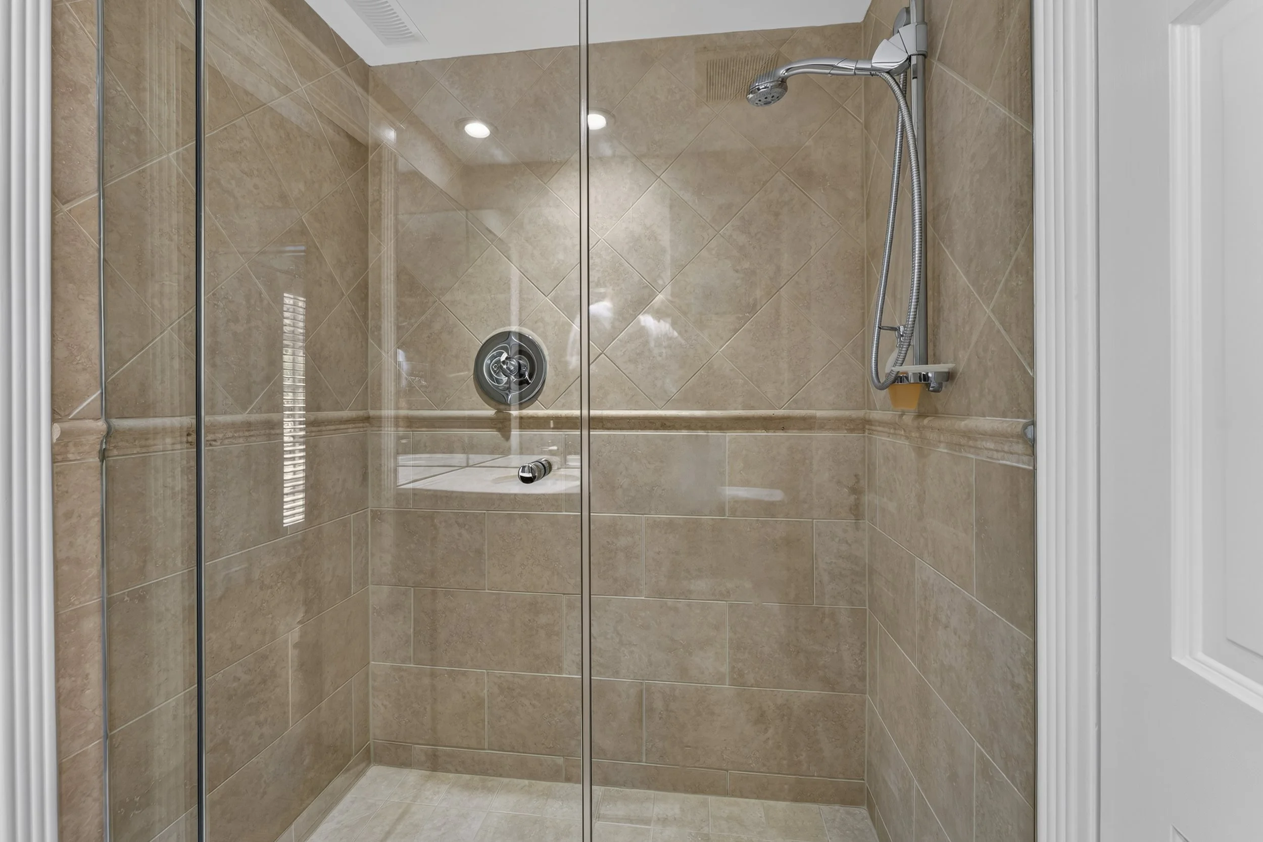 A glass-enclosed shower with beige tiled walls and a handheld showerhead. The shower has a glass door and a small built-in bench, with a vent or speaker on the ceiling.