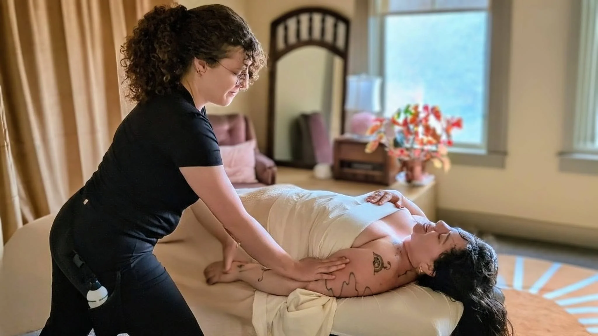 A person with curly hair and tattoos is receiving a massage from a massage therapist in a cozy, well-lit room with a window and a potted plant in the background.
