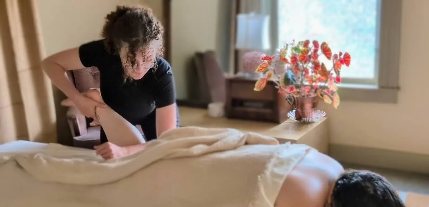 A woman is giving a massage to another person lying face down on a bed in a cozy room with a window and colorful flower arrangement on a side table.