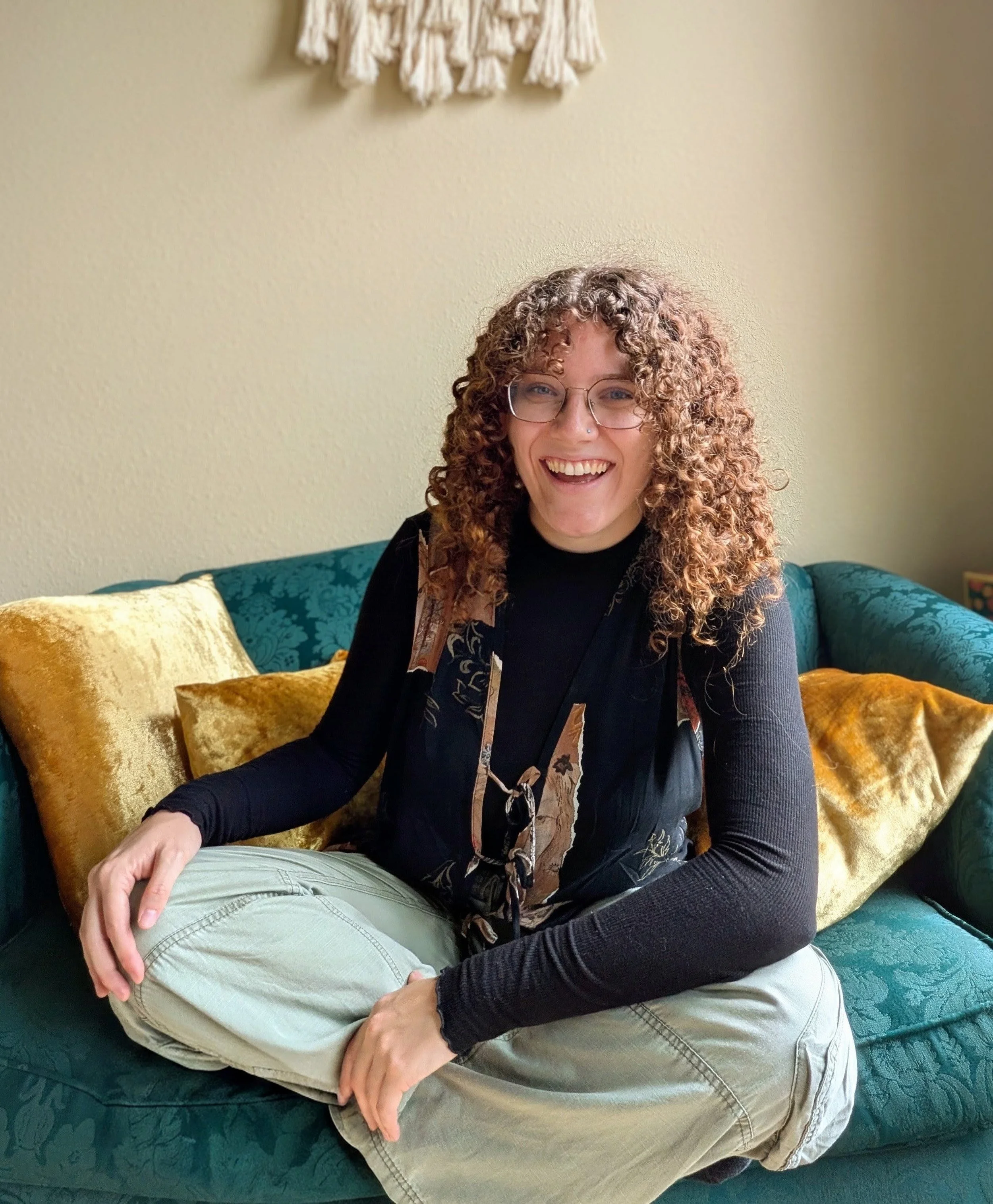 A woman with curly hair and glasses smiling while sitting on a teal couch with gold pillows, wearing a black top and light-colored pants.