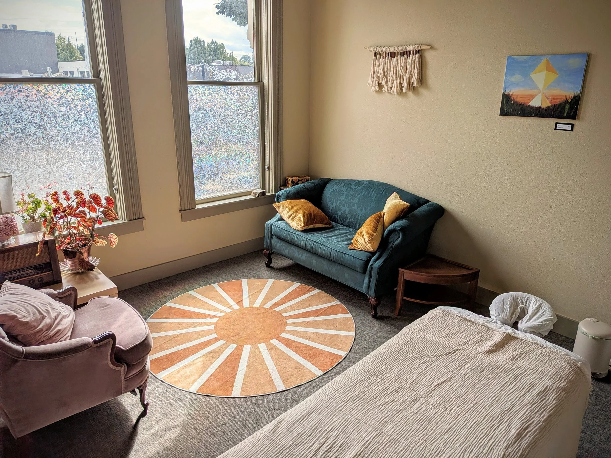A cozy living room with a vintage sofa, a floral armchair, and a colorful round rug. Two large windows with textured frosted glass allow natural light, and wall art hangs above a teal loveseat opposite a small wooden side table with a lamp.