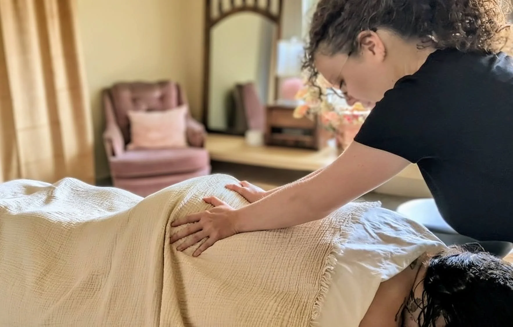 A person undergoing a massage, lying on a bed with a beige blanket, while another person applies pressure on their back in a cozy, well-decorated room with a pink armchair and floral decorations.
