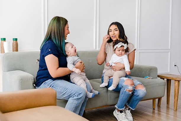 Two women sitting on a light gray couch with two young children. One woman with long green hair holding a baby, the other woman with a headband holding a toddler. The room has white walls and wooden furniture.